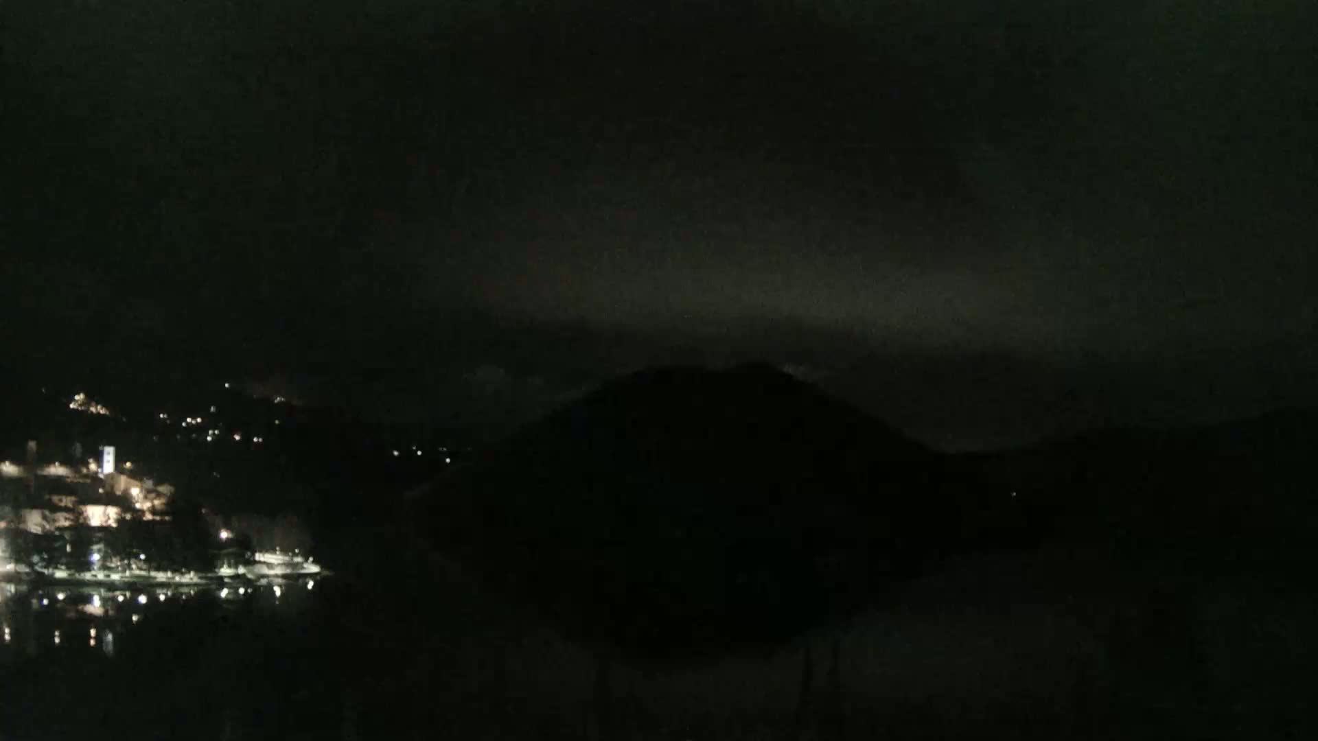 A nighttime, dark, and overcast view shows a body of water reflecting the faint lights of a town nestled among dark hills.