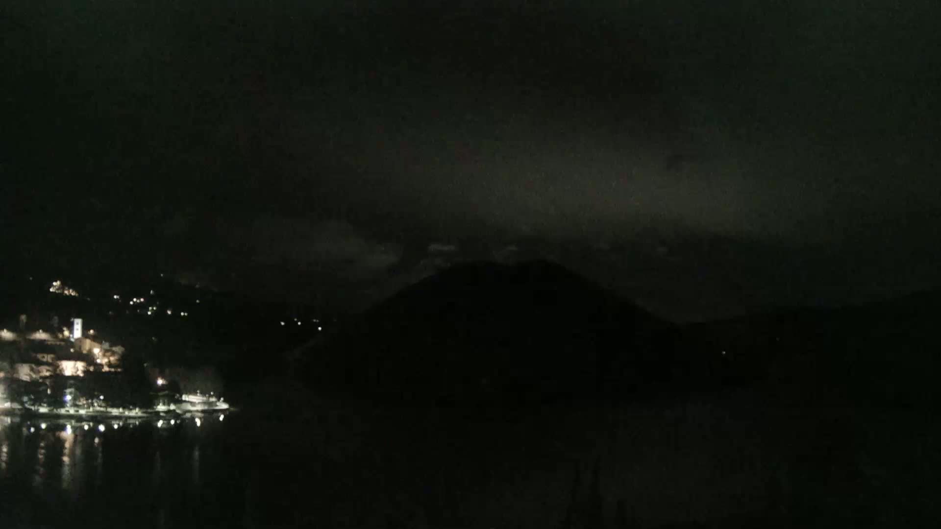 A dark, nighttime scene shows a body of water reflecting a few lights from a town on a hillside, with a large, dark hill or mountain in the middle ground under an overcast sky.