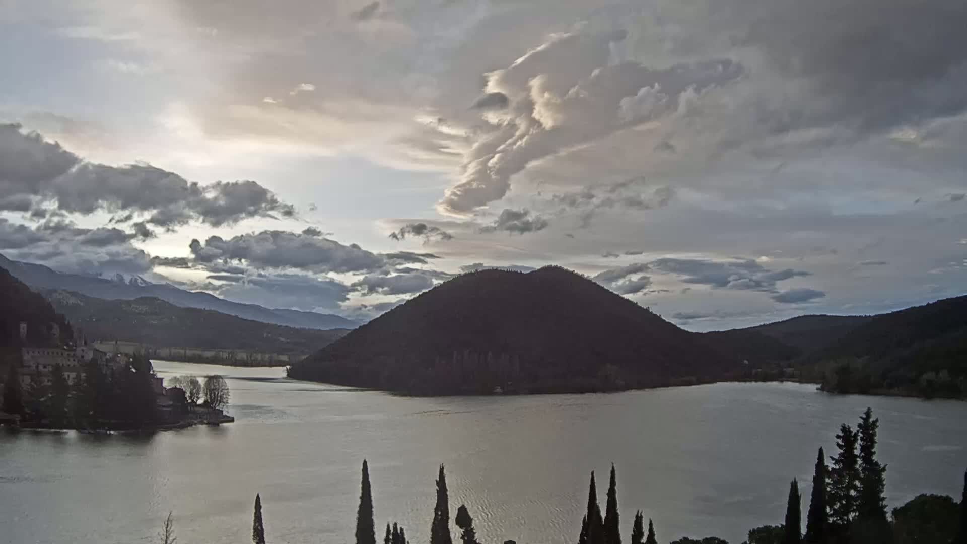 A calm lake is surrounded by hills and mountains under a cloudy sky.