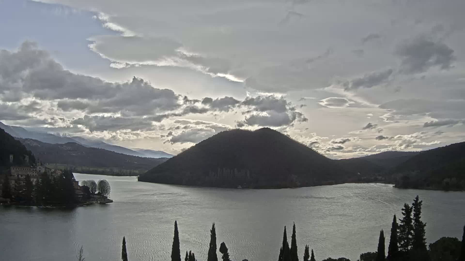 A lake is surrounded by hills and mountains under a cloudy sky.