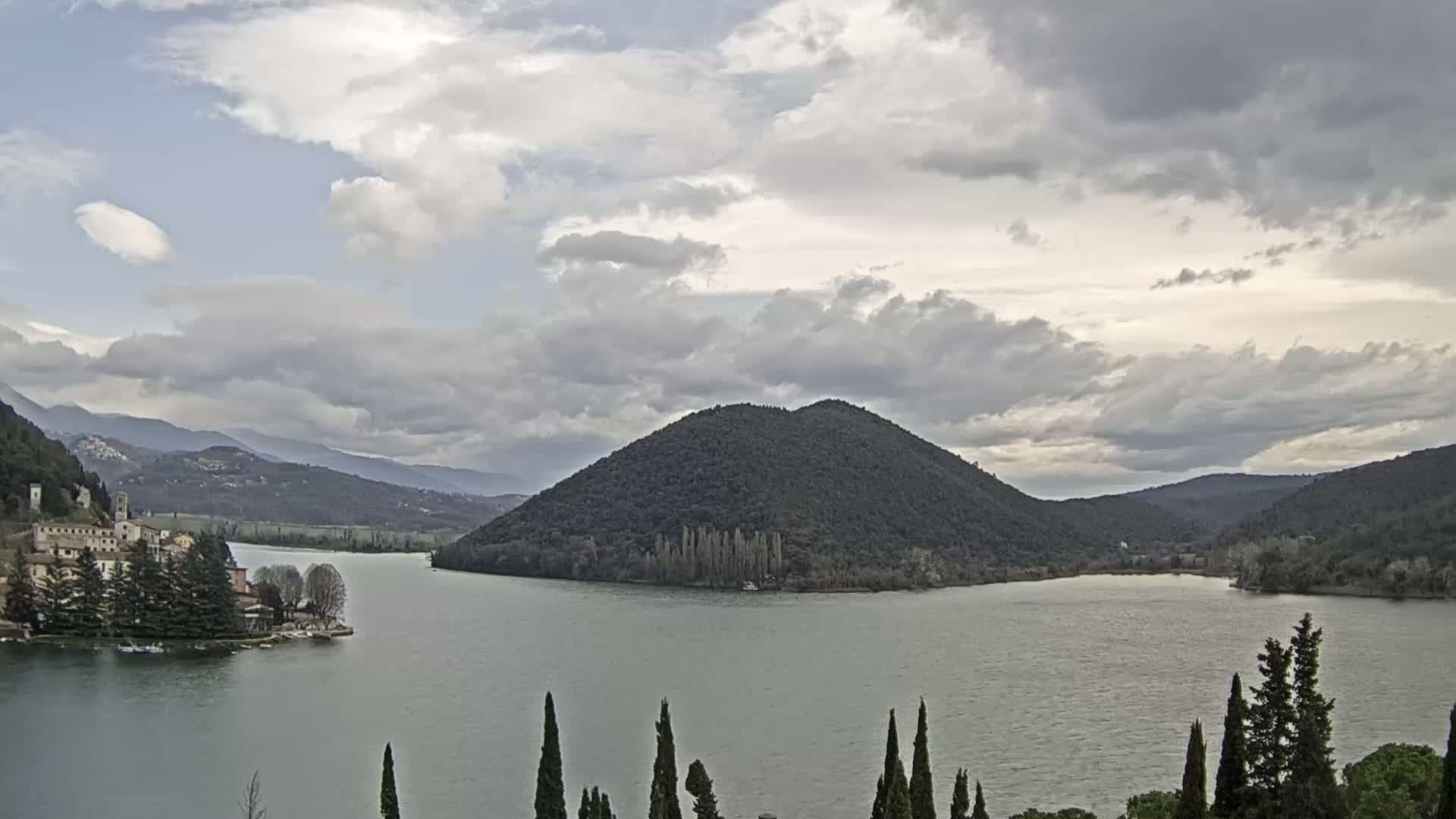 A lake is seen under a mostly cloudy sky, with a large, dark green island in the center and buildings and trees on a peninsula to the left, and mountains in the background.
