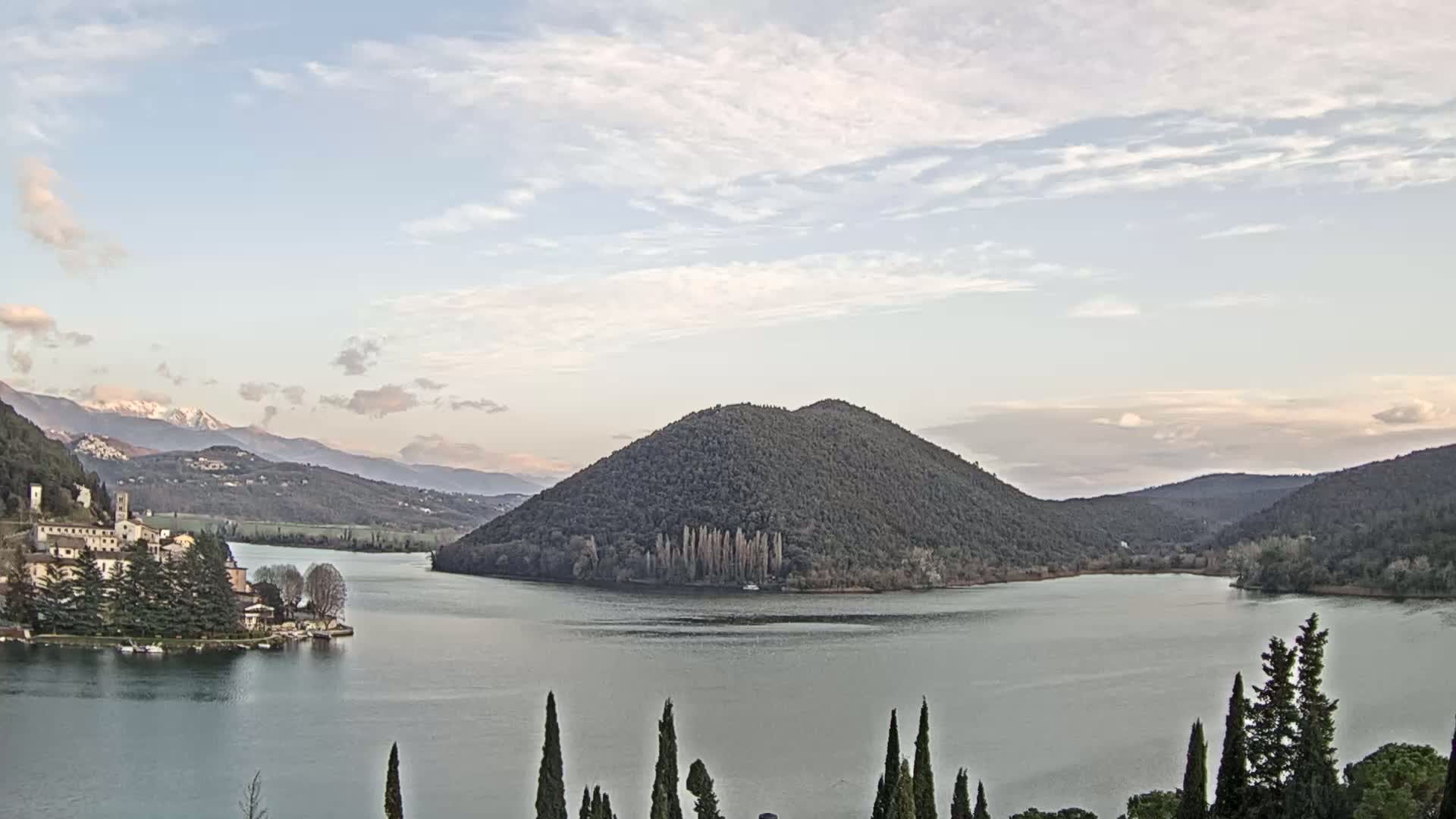 A calm lake is surrounded by a tree-covered hill, a village, and snow-capped mountains under a partly cloudy sky.