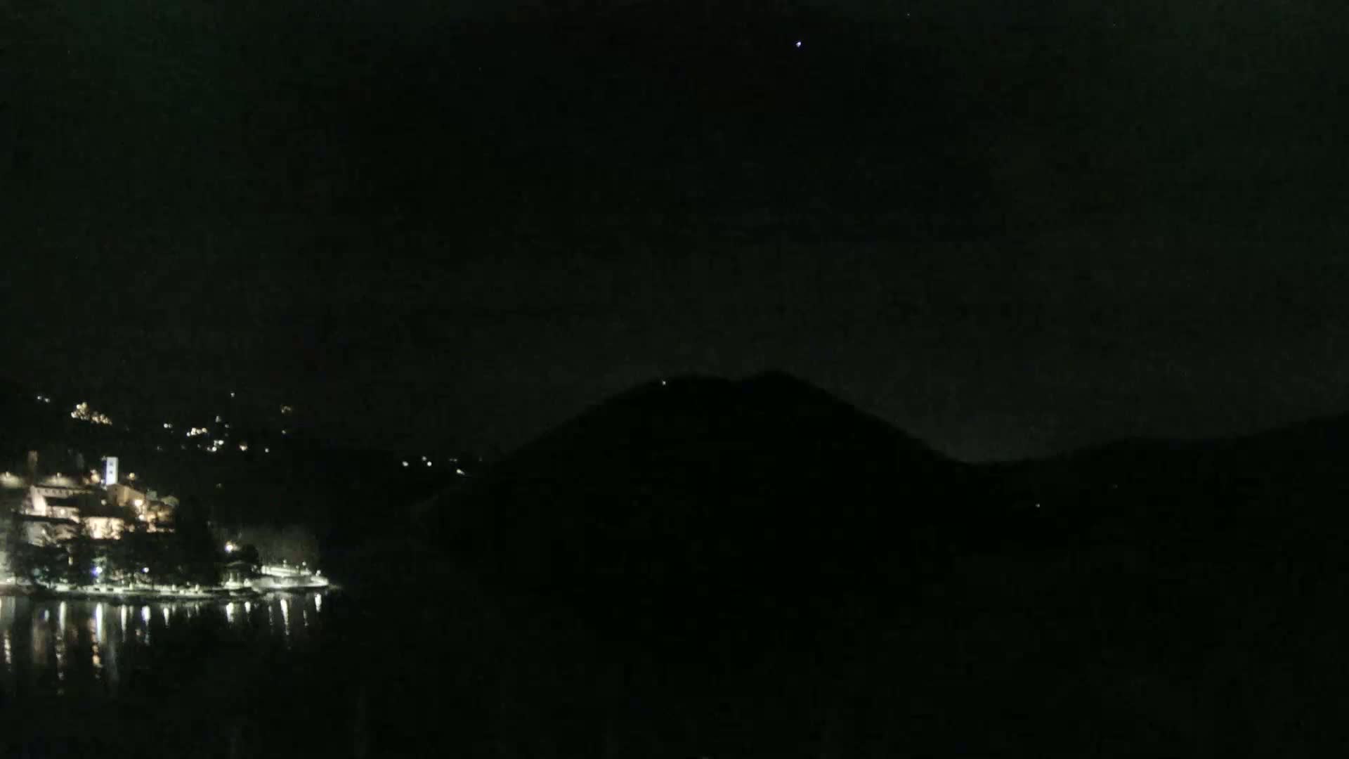 A nighttime view shows a dark mountain silhouetted against a clear, starlit sky overlooking a waterfront town with lights reflecting on the water.