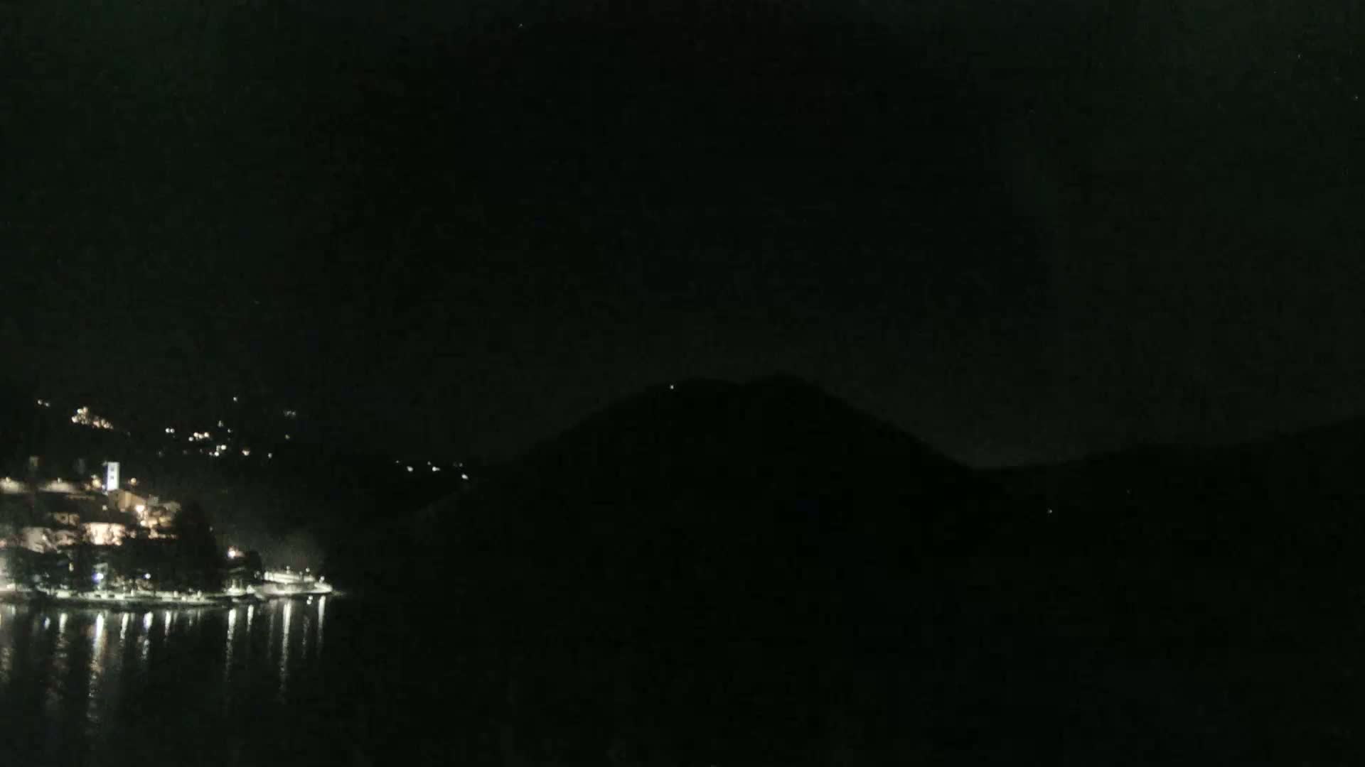 A dark nighttime scene shows a body of water reflecting the lights of a town nestled at the base of a hill.