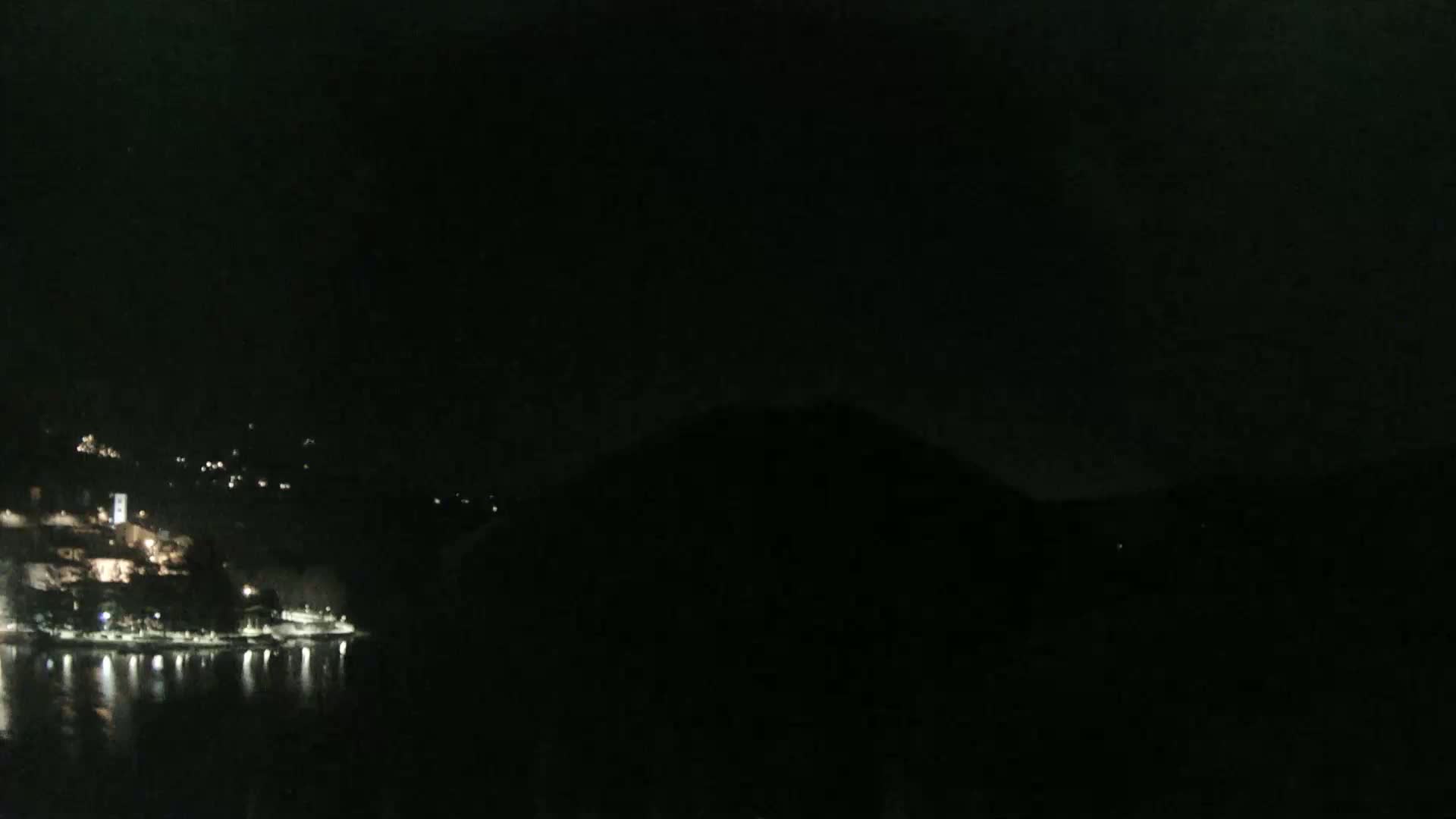 A dark nighttime scene shows a dimly lit waterfront town nestled against a hill, with calm water reflecting the lights.