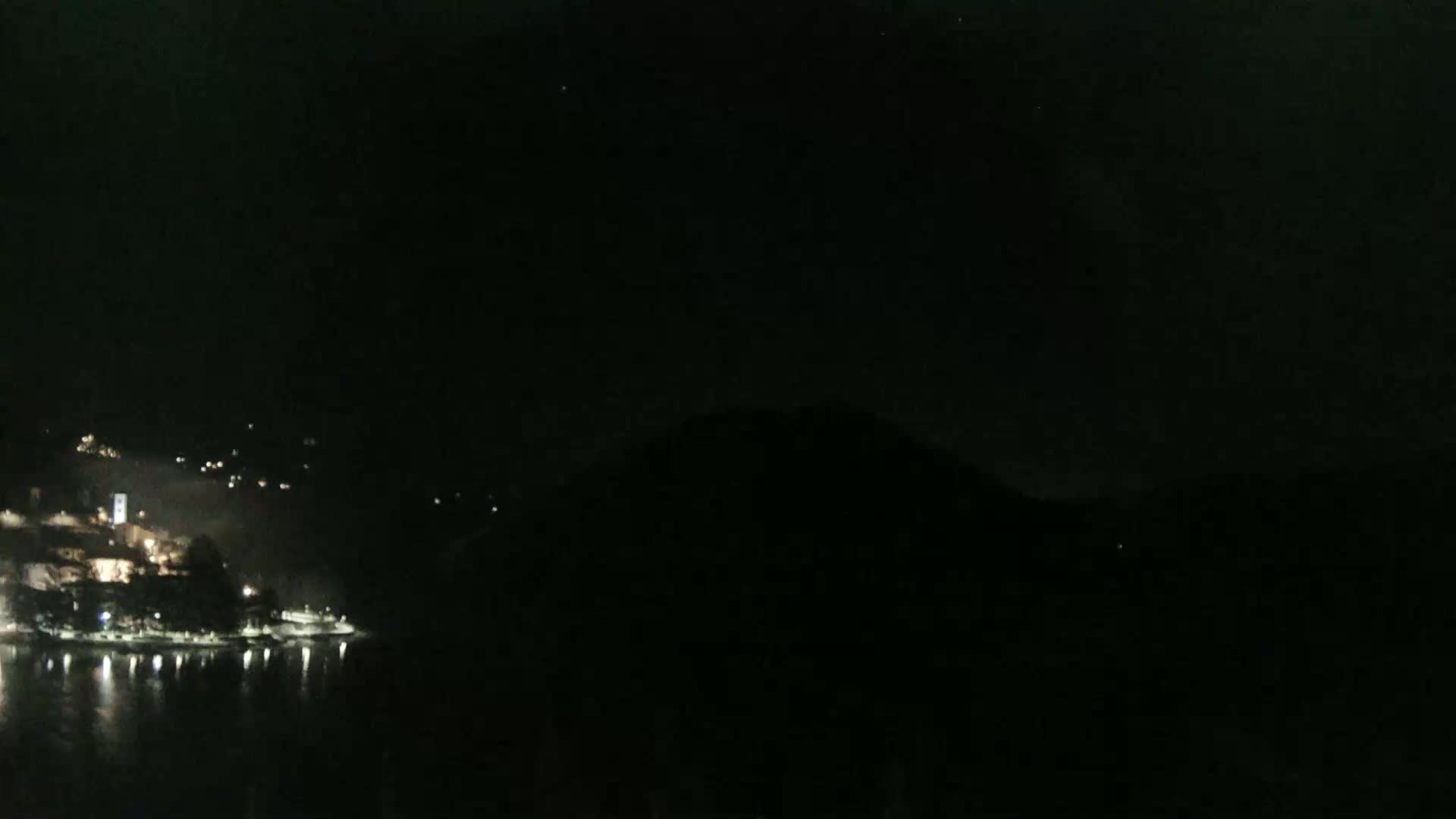 A dark night scene shows a body of water reflecting the lights of a town nestled against a hill.