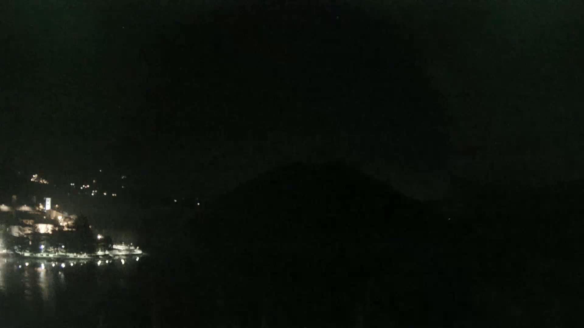 A nighttime view shows a dark body of water with a dimly lit town on the opposite shore, nestled against a dark hill under a mostly clear sky.