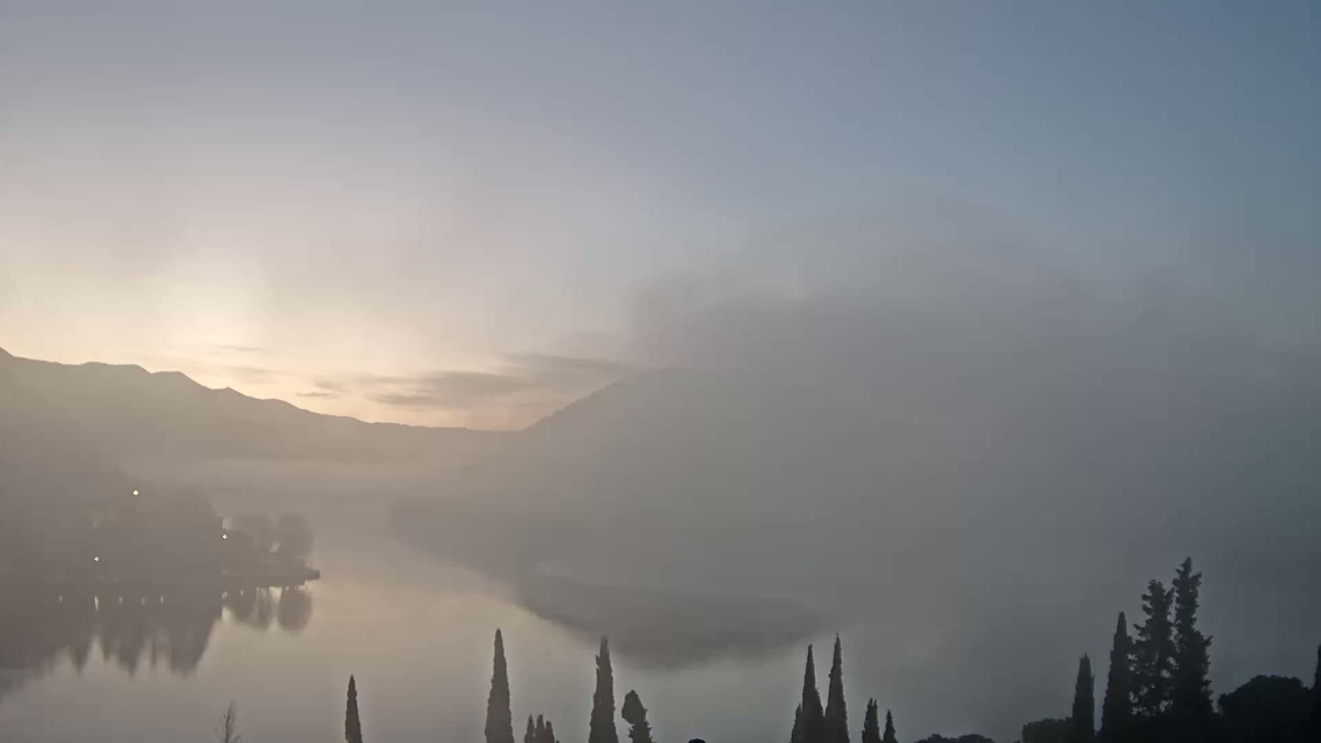 A calm lake is shrouded in morning fog, with mountains in the background and silhouetted trees in the foreground under a pale sunrise.