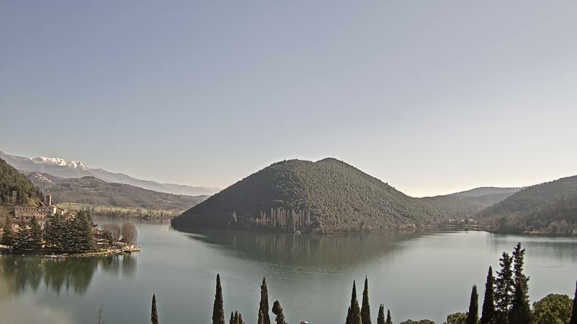 A tranquil lake scene under a clear sky shows a village nestled on its shore, a tree-covered hill rising from the water, and snow-capped mountains in the distance.