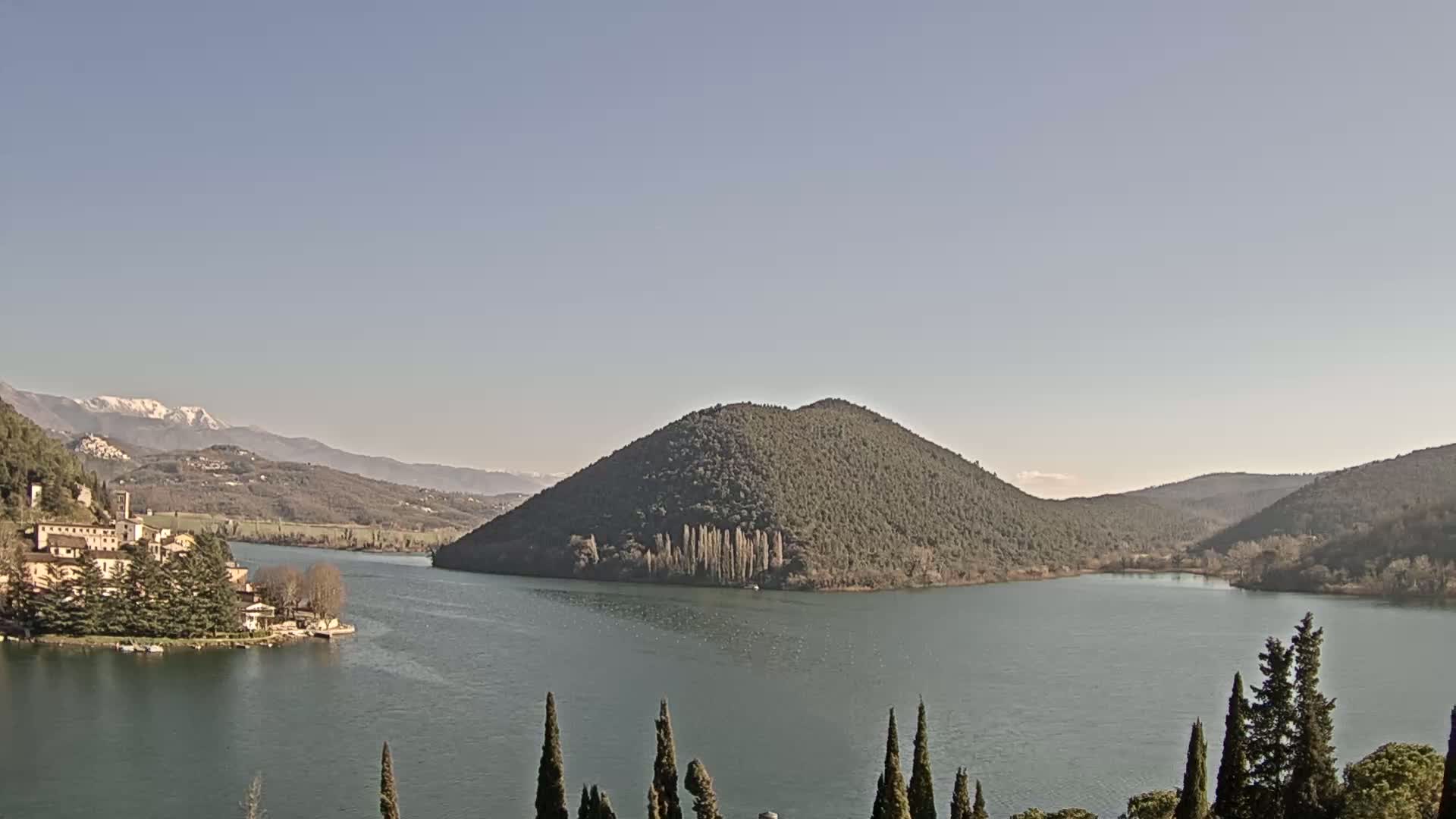 A lake, nestled between tree-covered hills and mountains with snow-capped peaks in the distance, is visible under a clear, sunny sky.