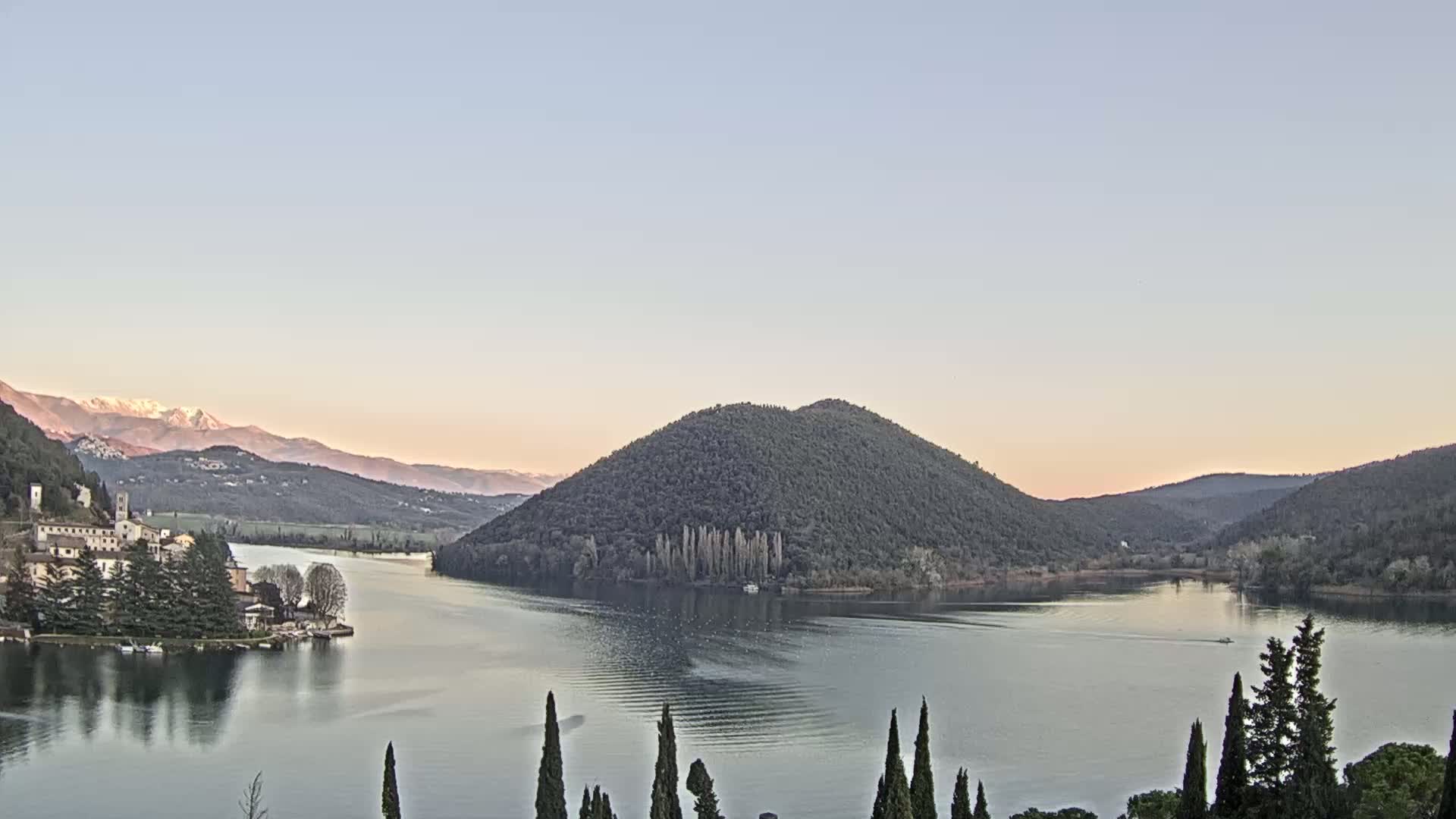 A tranquil lake scene at sunset features a village nestled on a lakeshore, a prominent forested hill in the center, and snow-capped mountains in the background under a clear, pale sky.