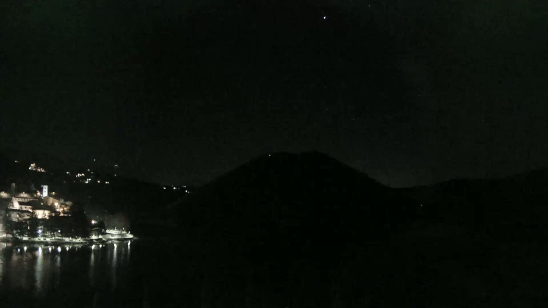 A dark night scene shows a body of water reflecting scattered lights from a town nestled on a hillside, with a large, dark hill silhouetted against the starry sky.