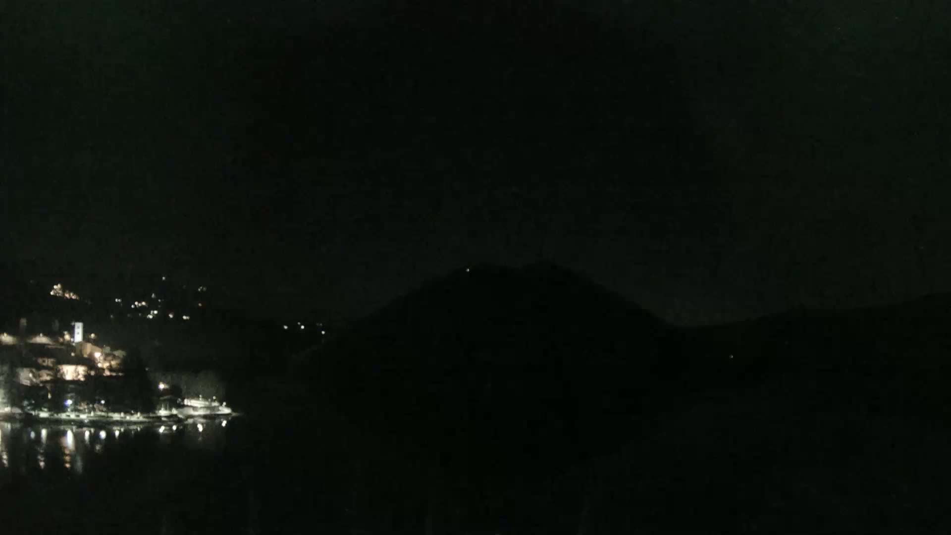 A nighttime view shows a dark body of water reflecting scattered lights from a town nestled on a hillside, with a dark mountain looming in the background under a clear, dark sky.
