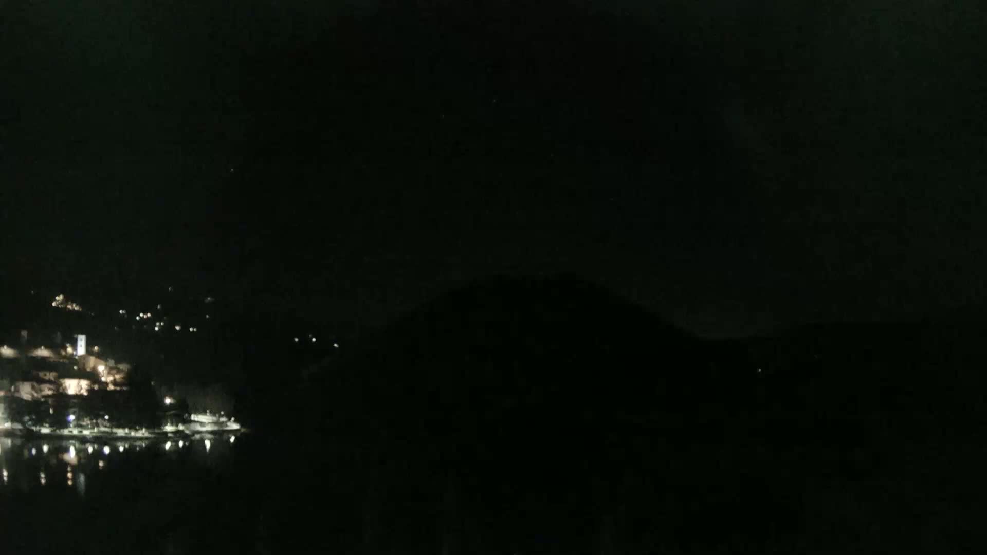 A dark, nighttime scene shows a dimly lit waterfront town nestled between hills, reflected in a calm body of water.