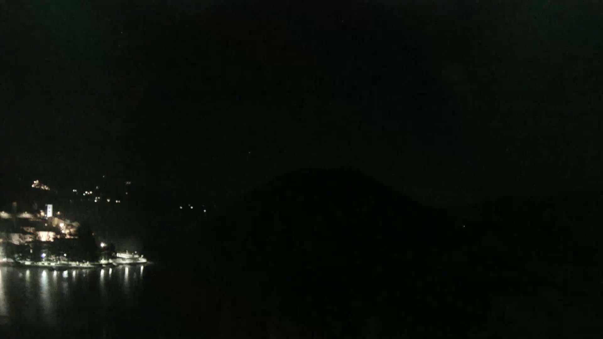 A nighttime view of a town's lights along a dark body of water, bordered by hills under a clear, dark sky.