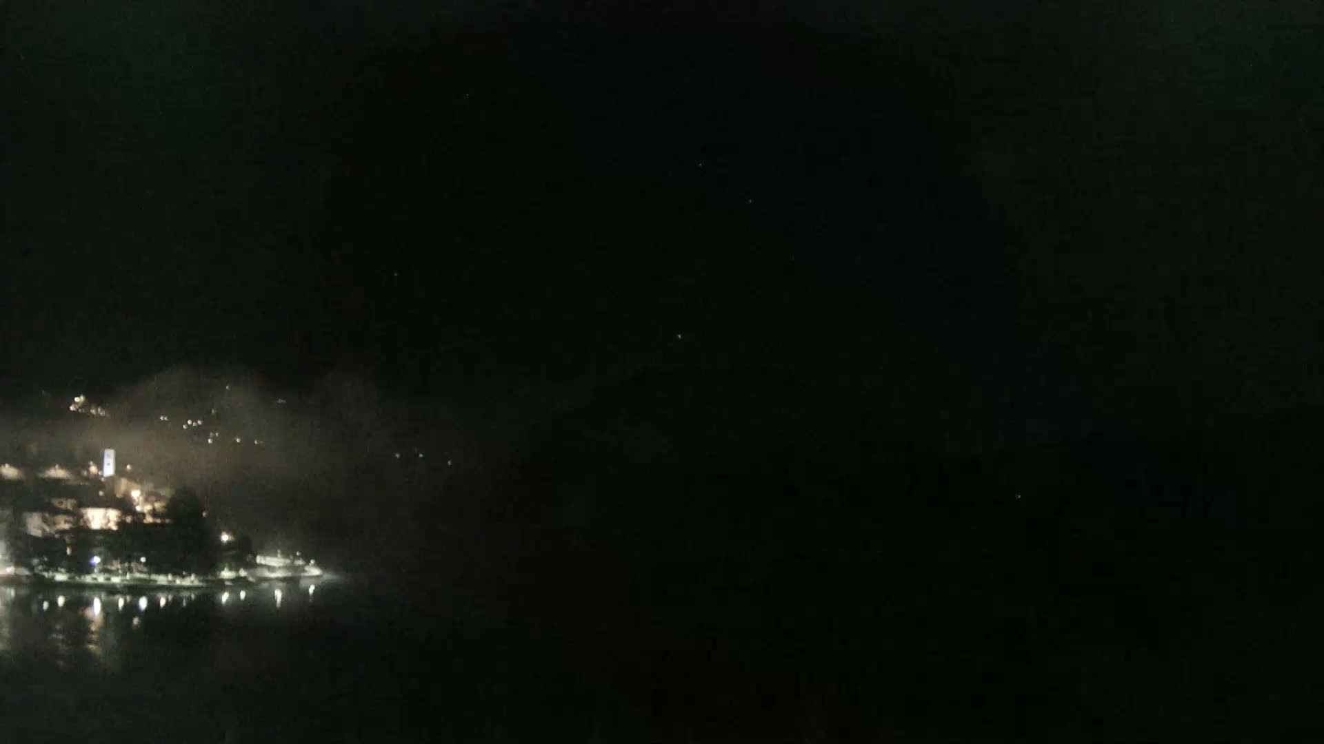 A nighttime view shows a coastal town with faint lights partially obscured by fog or low clouds over a dark body of water.