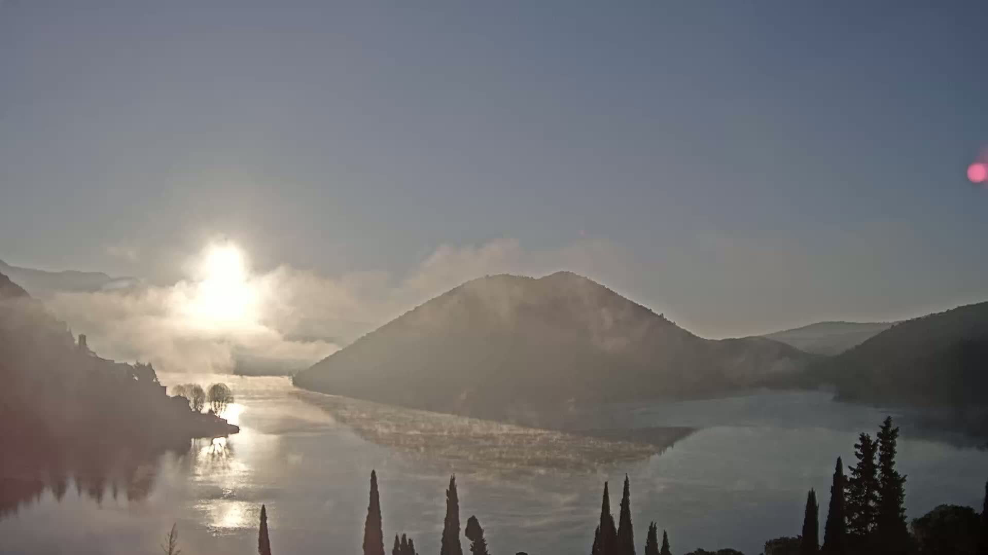 A sunlit, misty sunrise over a calm lake nestled between rolling hills.