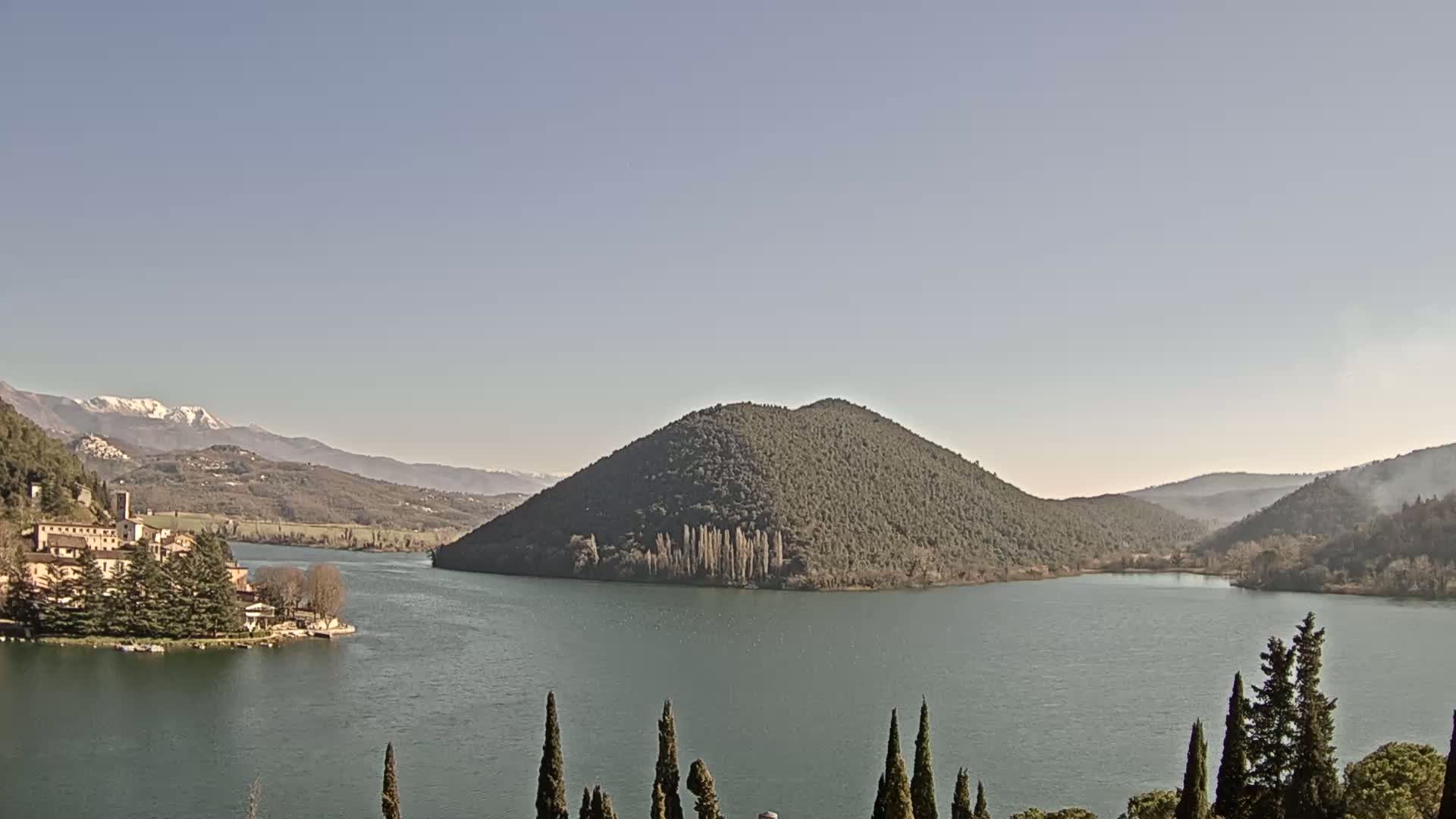 A lake surrounded by mountains, one of which is a rounded, forested hill, with a village and snow-capped mountains in the background under a clear sky.