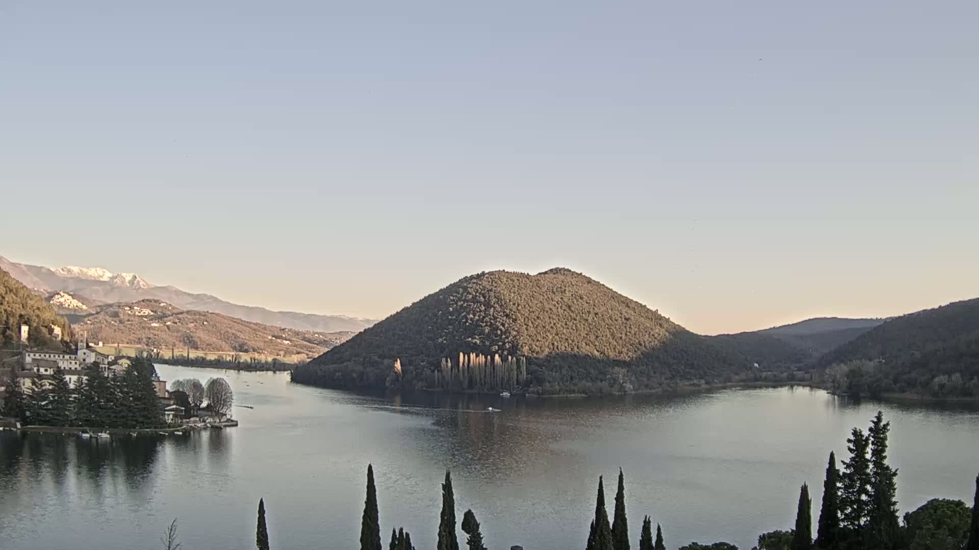 A calm lake nestled in a valley between rolling hills, with snow-capped mountains in the background under a clear sky.