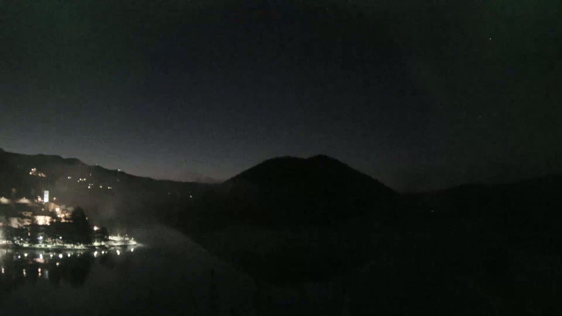 A dark, nighttime scene shows a body of water reflecting the lights of a town nestled between hills under a mostly clear, starlit sky.