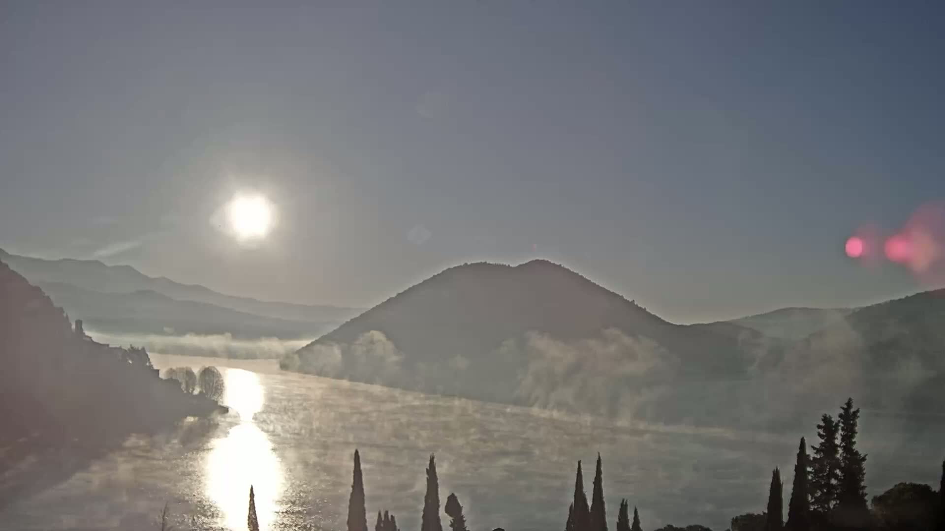 A sunlit river valley is shrouded in morning mist, with mountains rising on either side and dark evergreens in the foreground.