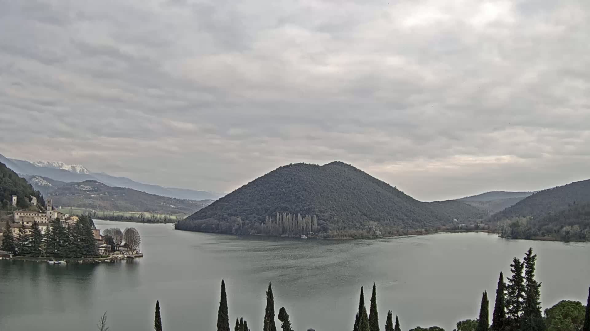 A lake is surrounded by hills and mountains under a cloudy sky, with a village visible on a peninsula in the foreground.