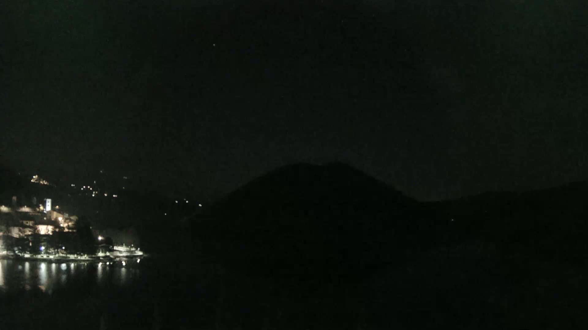 A nighttime view shows a dark, hilly landscape with a body of water reflecting scattered lights from a town on the opposite shore under a clear, starlit sky.