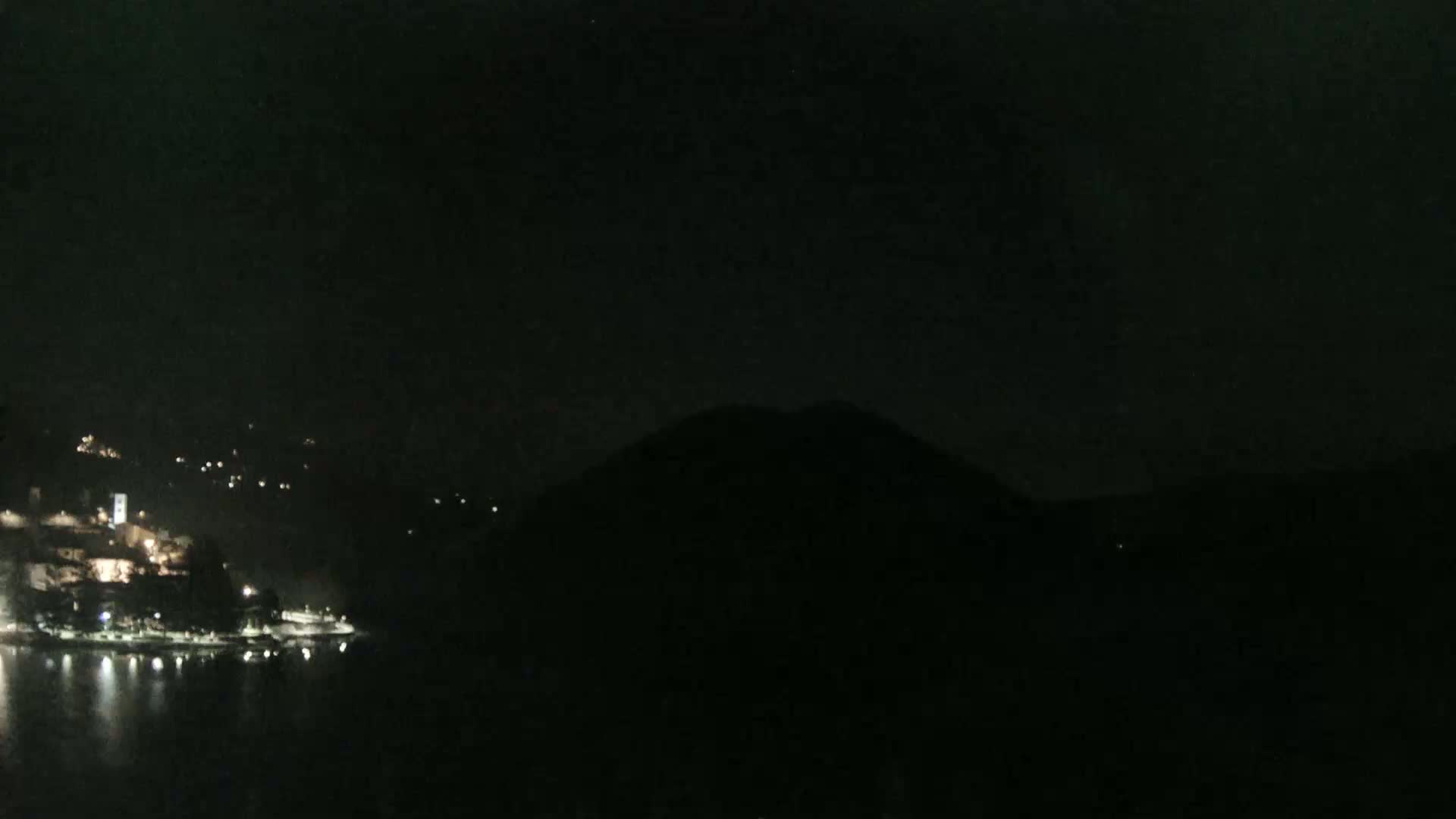 A dark, nighttime scene shows a body of water reflecting the lights of a small town nestled on a hillside, with a large, silhouetted hill in the foreground.