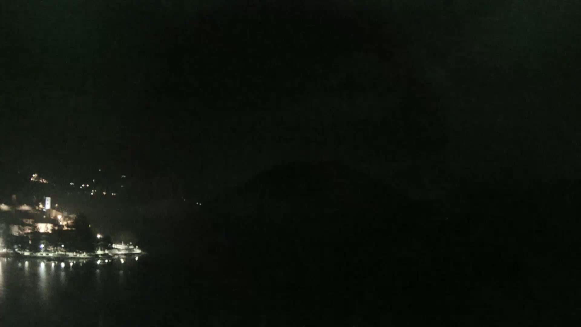 A nighttime view of a dark, calm body of water reflecting the faint lights of a town nestled against a hillside.