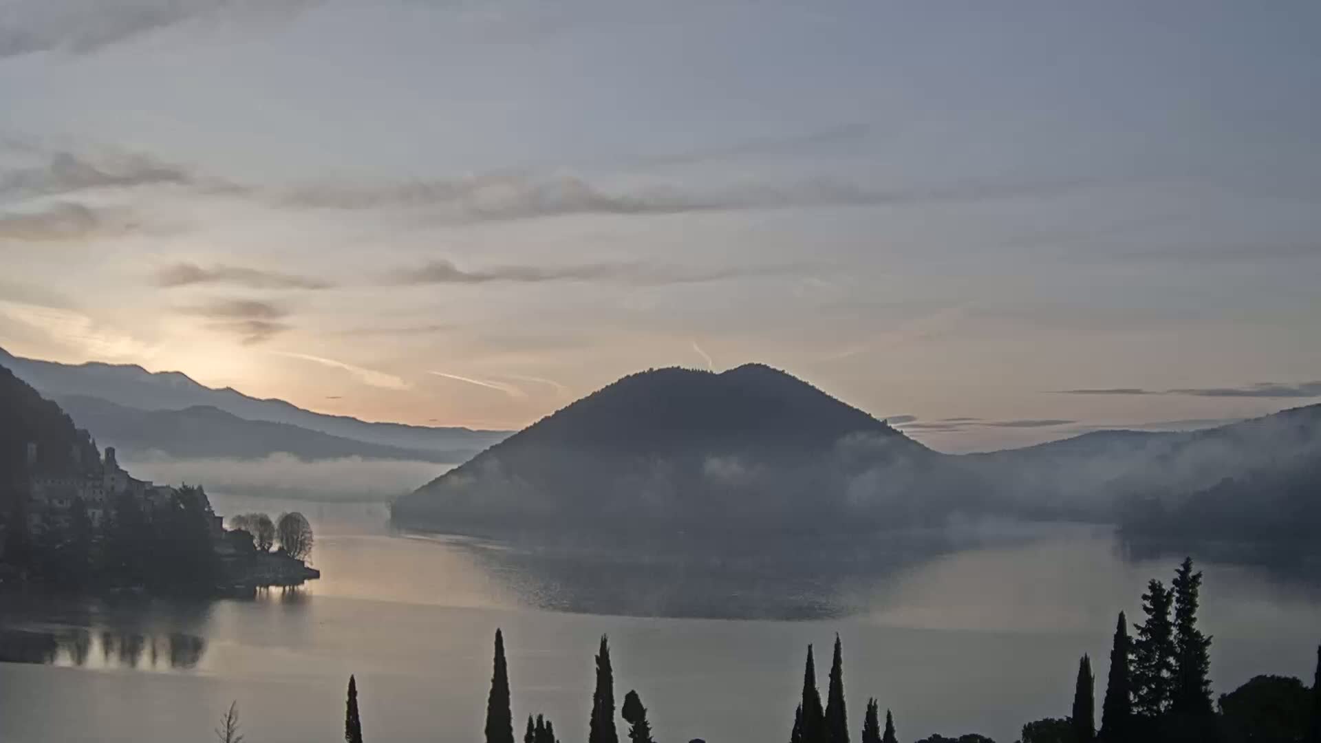 A calm lake at sunrise is shrouded in mist, with a prominent hill in the center and mountains in the background.