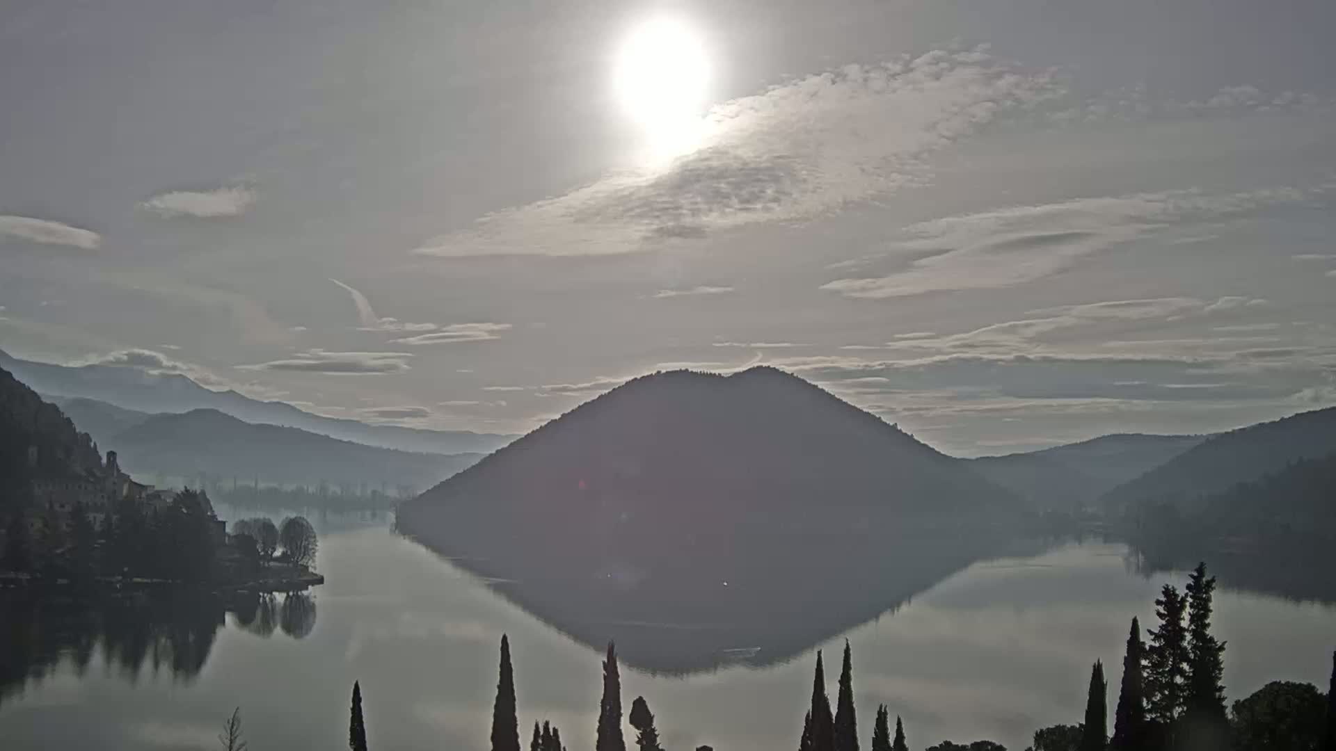 A calm lake reflects a sunlit sky and a hazy, mountainous landscape under a mostly cloudy sky.