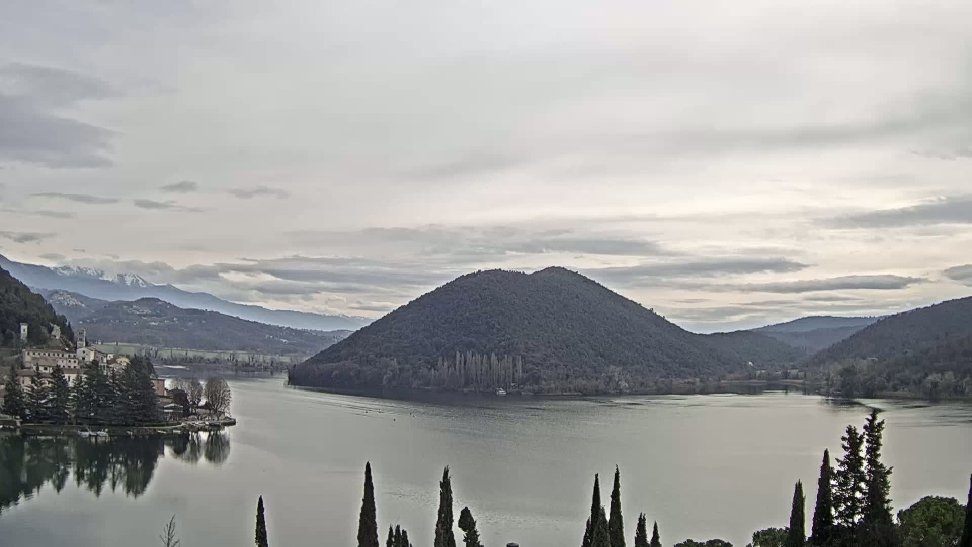 A calm lake is nestled among rolling hills and mountains under a cloudy sky, with a snow-capped mountain range visible in the distance.