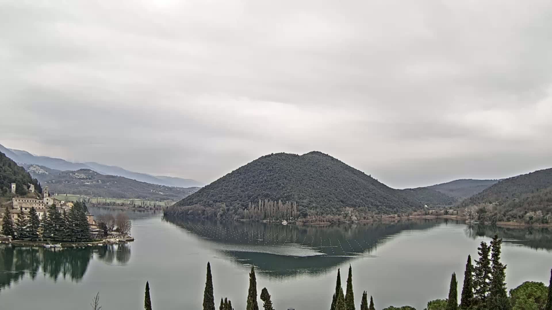 A calm lake is surrounded by tree-covered hills and mountains under an overcast sky.