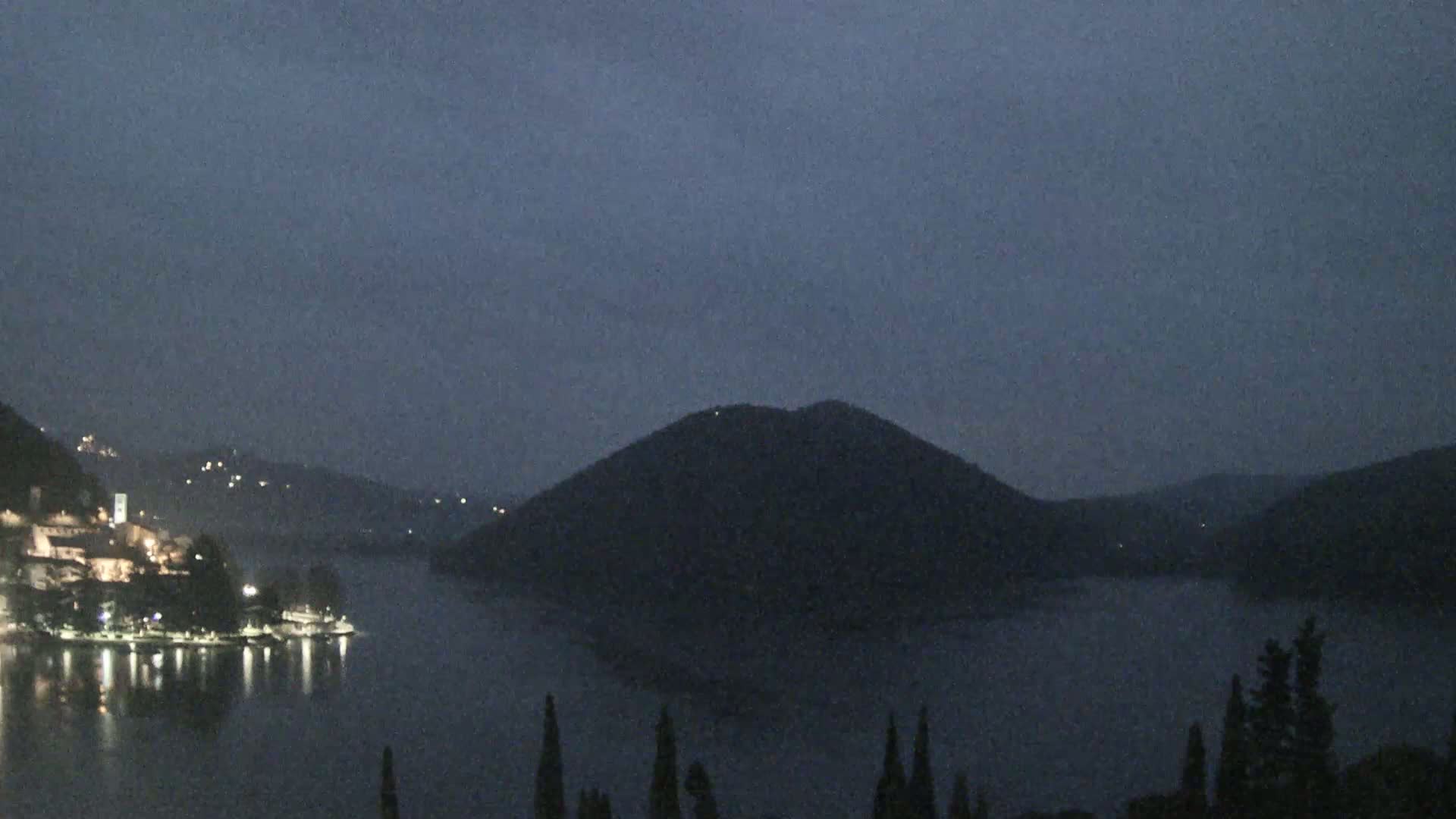 A nighttime view of a lake, with a town's lights visible on one shore and a large, dark hill dominating the opposite shore under a dusky sky.