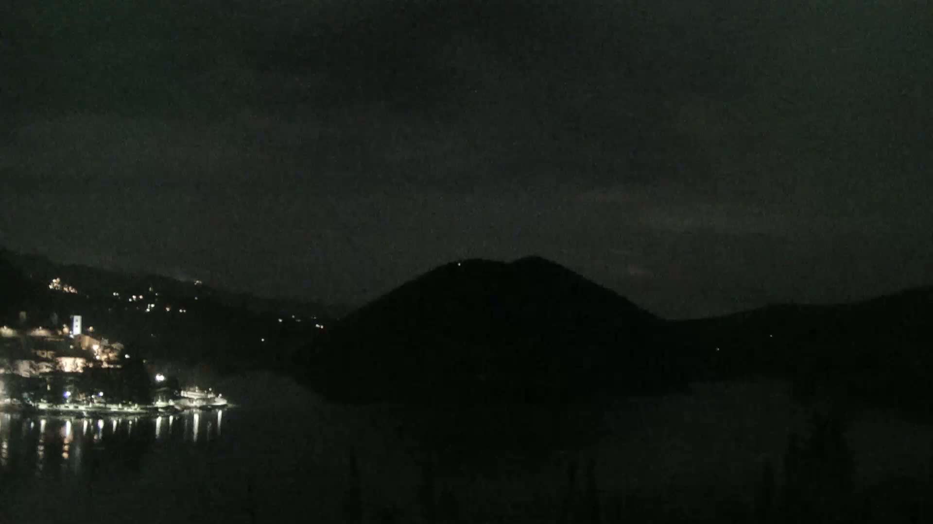 A dark night scene shows a body of water reflecting the lights of a town nestled at the base of a hill, under a mostly cloudy sky.