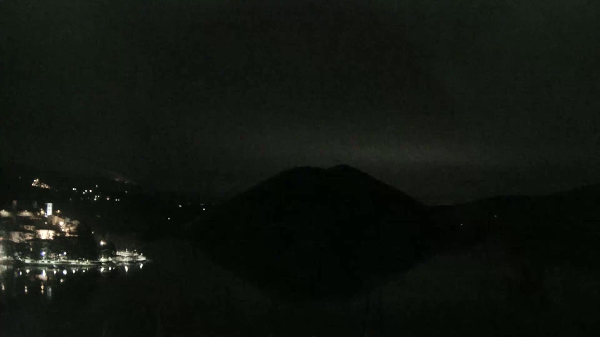A dark, nighttime scene shows a body of water reflecting a few lights from a town nestled in hills across from a silhouetted mountain under a mostly cloudy sky.