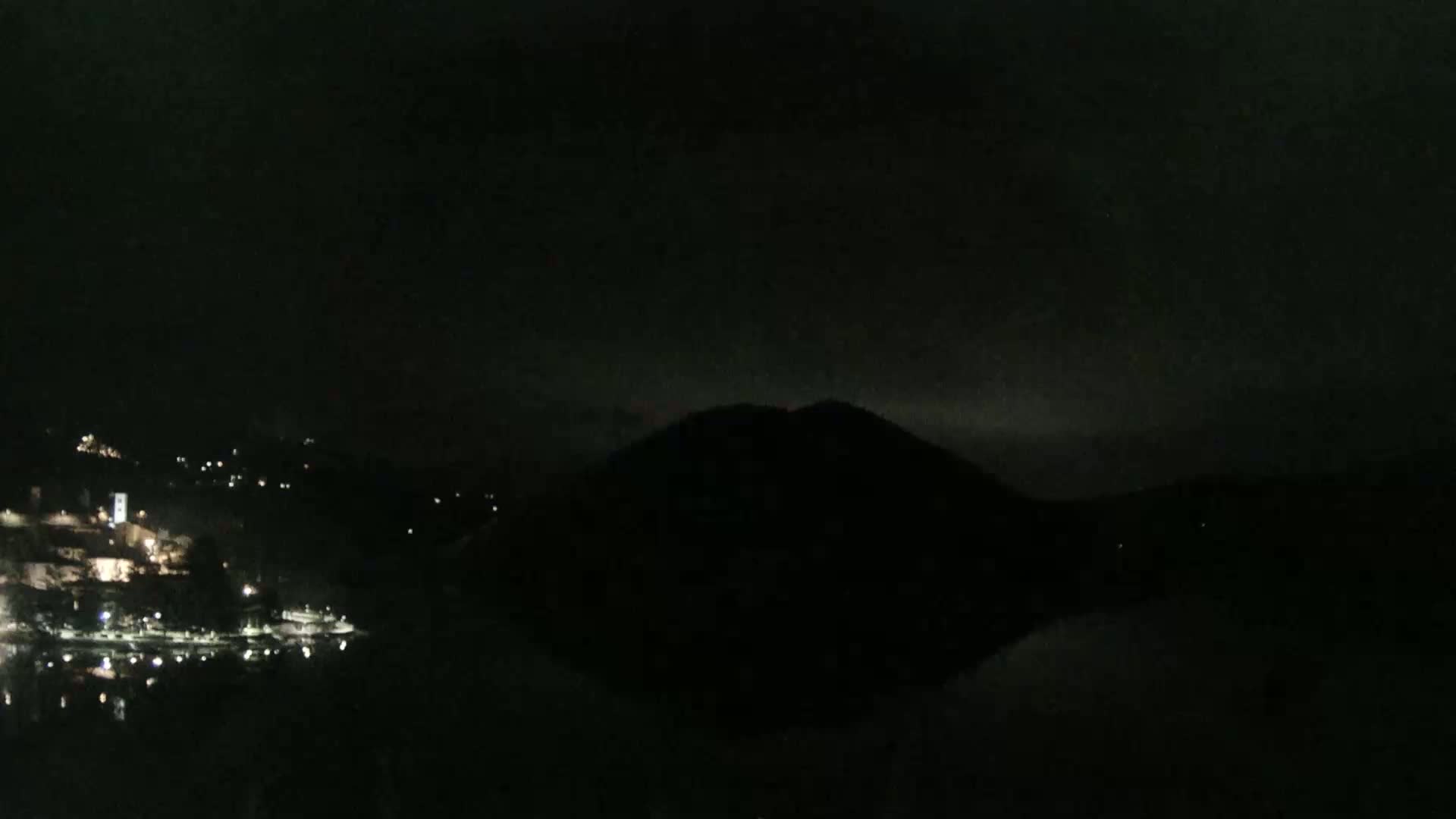 A dark, nighttime scene shows a body of water reflecting a silhouetted hill, with faintly visible lights from a town on the opposite shore.