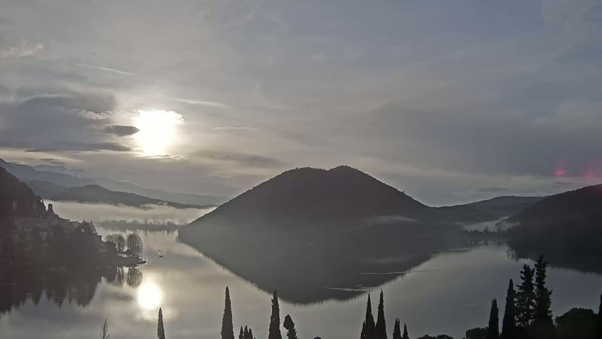 A calm lake reflecting a hazy sunrise, with mist covering parts of the surrounding hills and mountains.