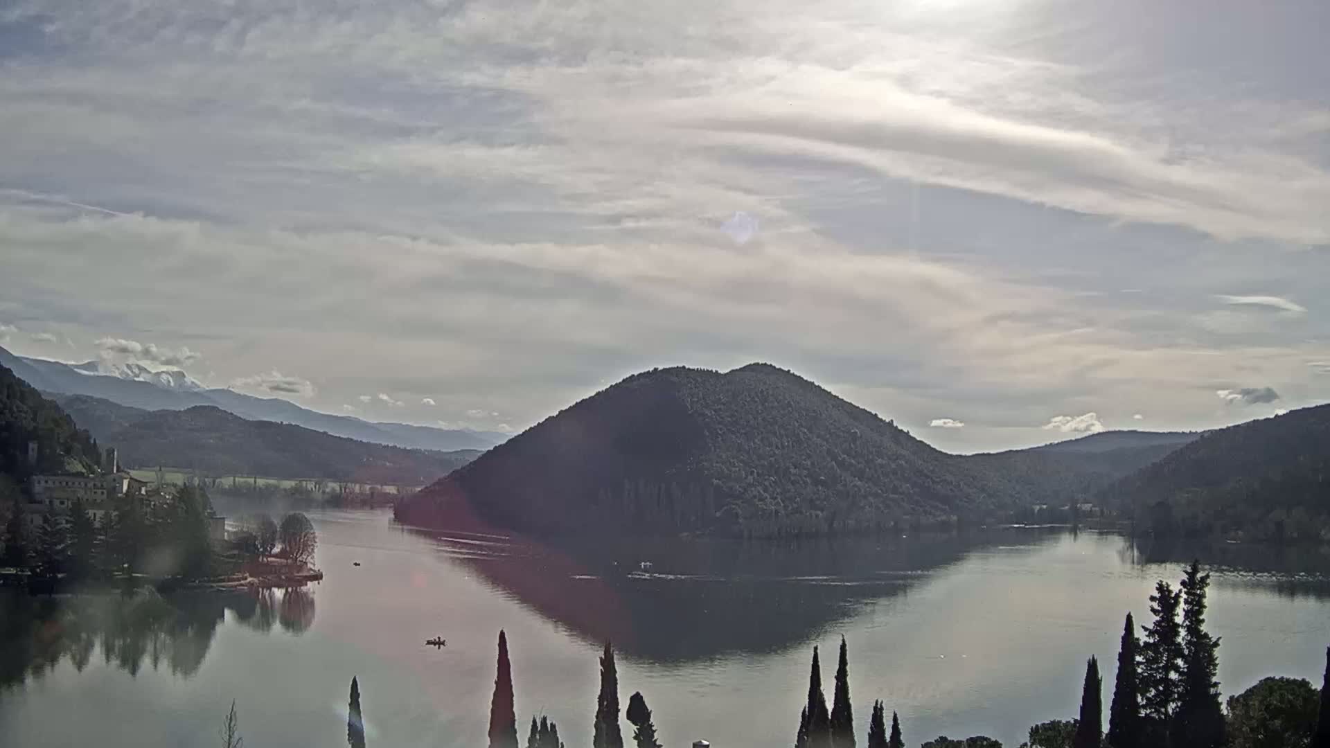 A calm lake reflects a conical island and distant mountains under a mostly cloudy sky.