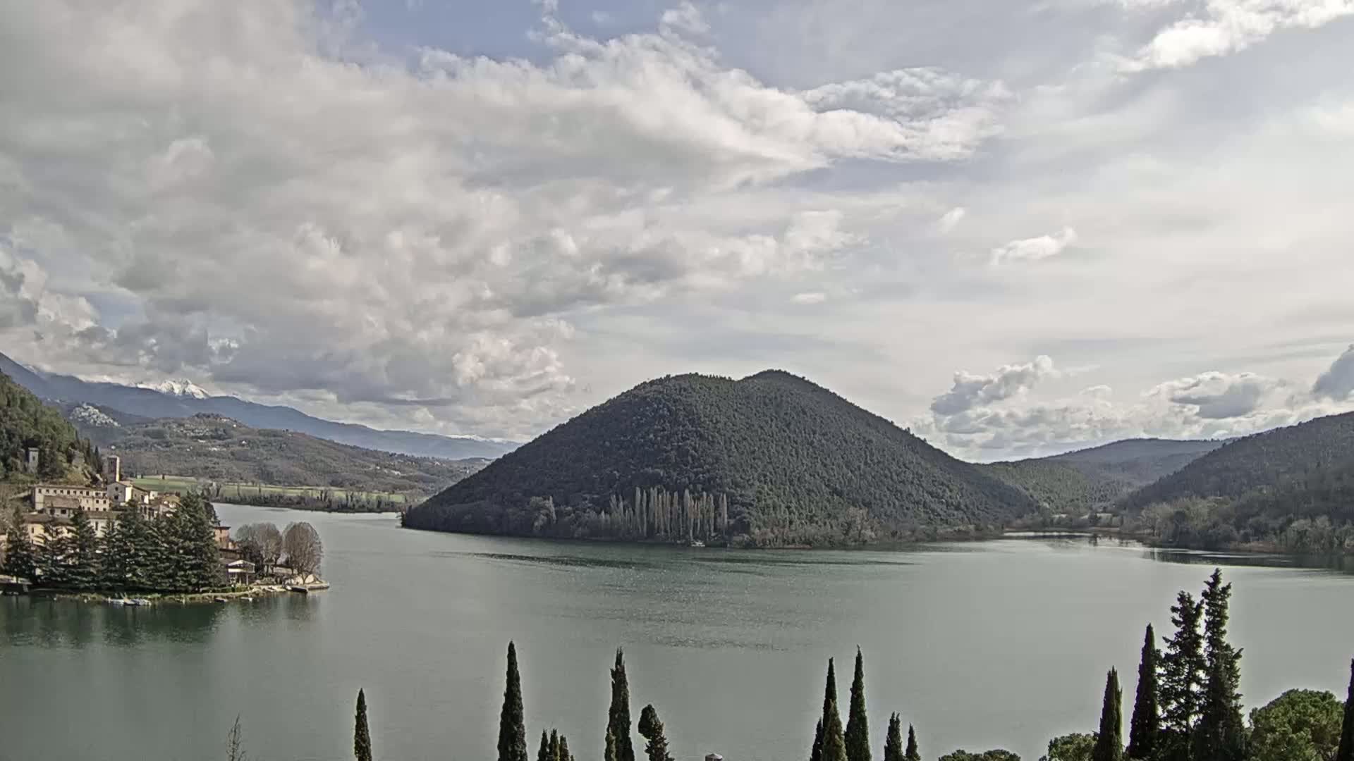 A lake is surrounded by hills and mountains under a partly cloudy sky, with a village visible on the left shore.