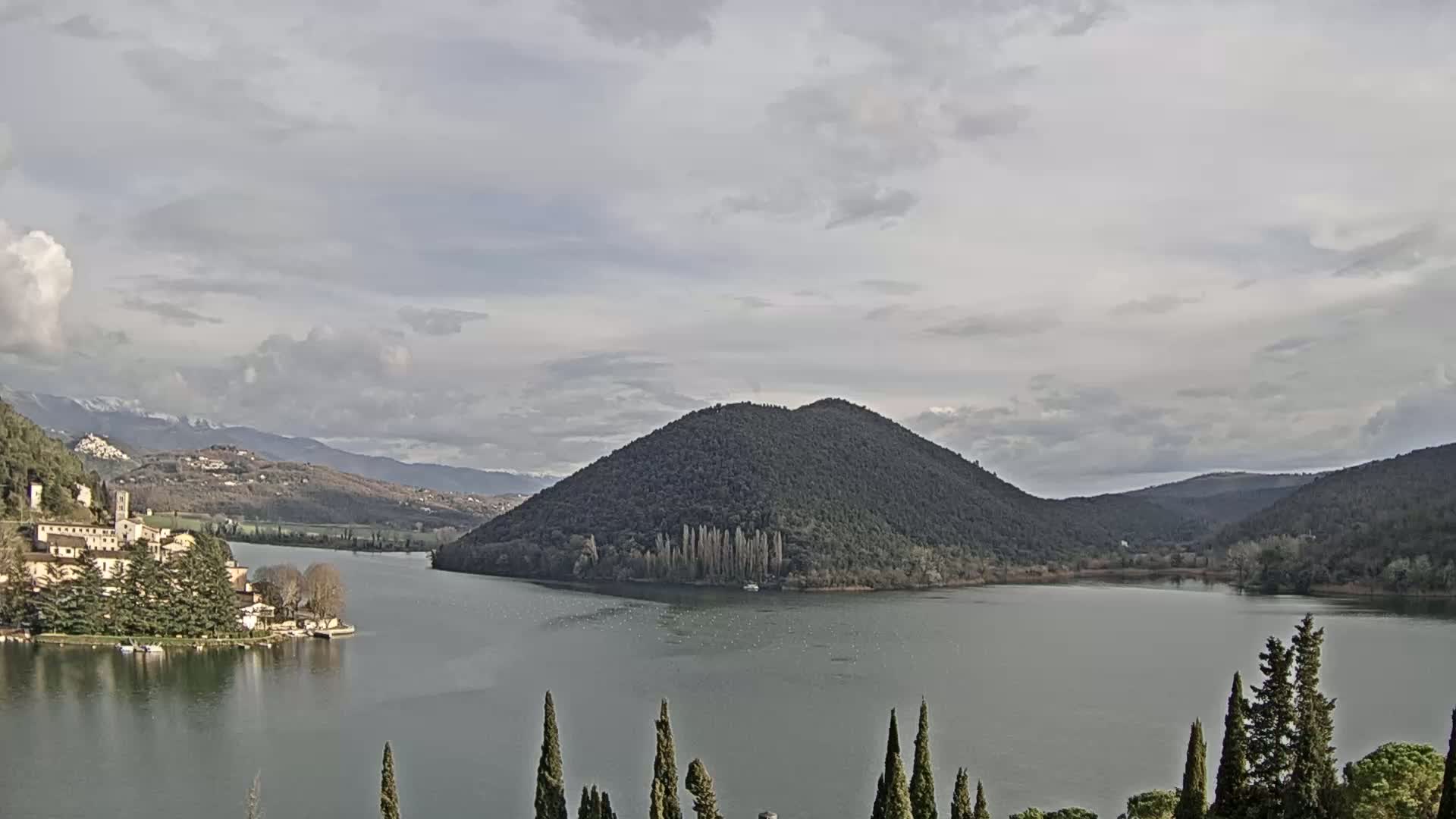 A calm lake is surrounded by hills and mountains under a cloudy sky, with a village and a distinctive, dark, cone-shaped hill visible in the mid-ground.