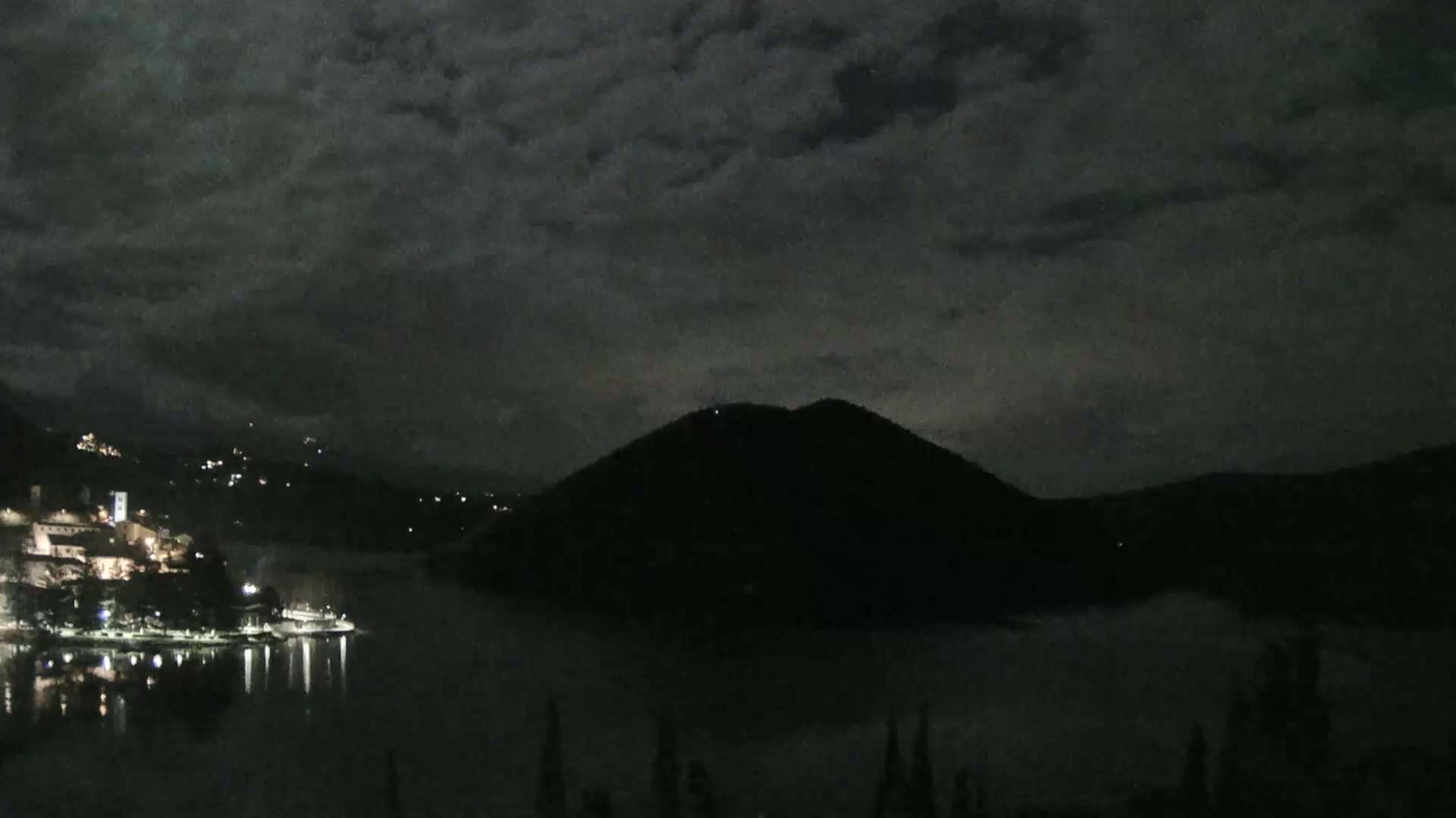 A nighttime view of a town and lake nestled between dark hills under a cloudy sky.