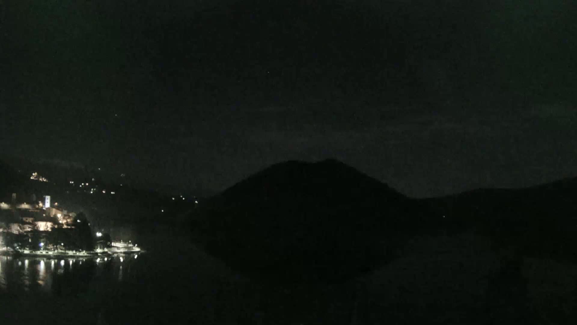 A dark, nighttime scene shows a body of water reflecting the lights of a town nestled on a hillside across from a large, dark mountain under a mostly clear sky.