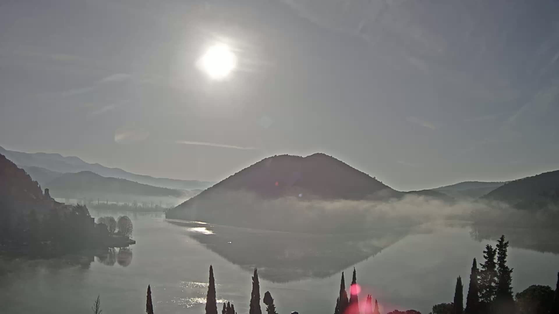 A calm lake is partially covered in fog at sunrise, with mountains and trees in the background.