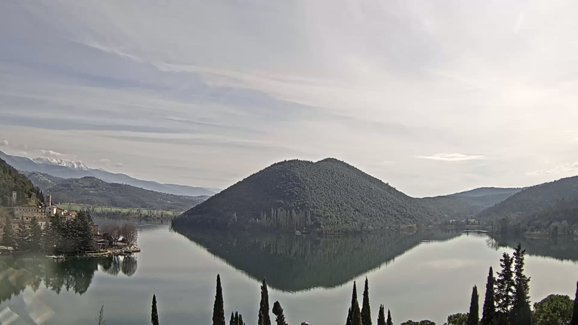 A calm lake reflects a tree-covered hill and distant snow-capped mountains under a mostly cloudy sky.