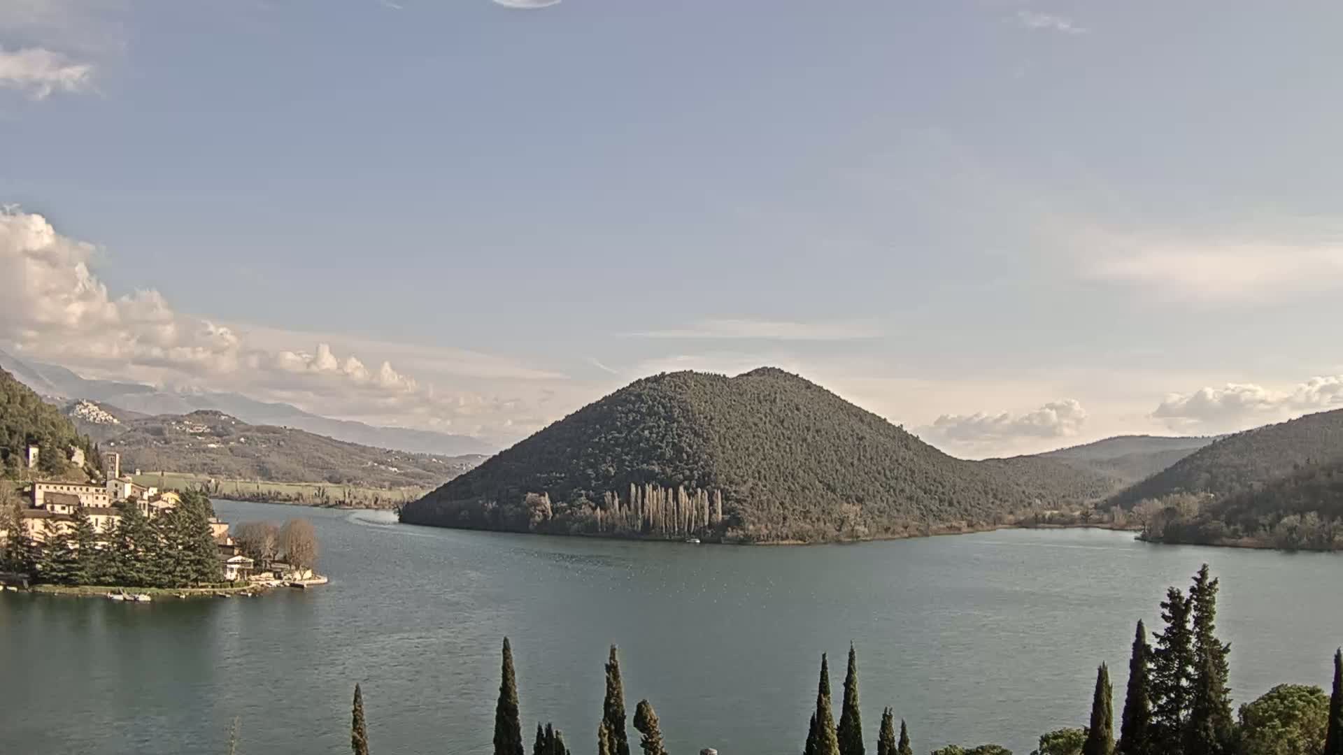 A lake is nestled between rolling hills under a mostly clear sky with scattered clouds, a village situated on the lakeshore to the left.