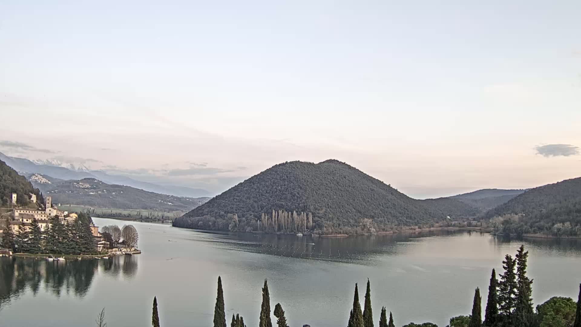 A calm lake reflects a village and forested hills under a pale, clear sky with distant snow-capped mountains.