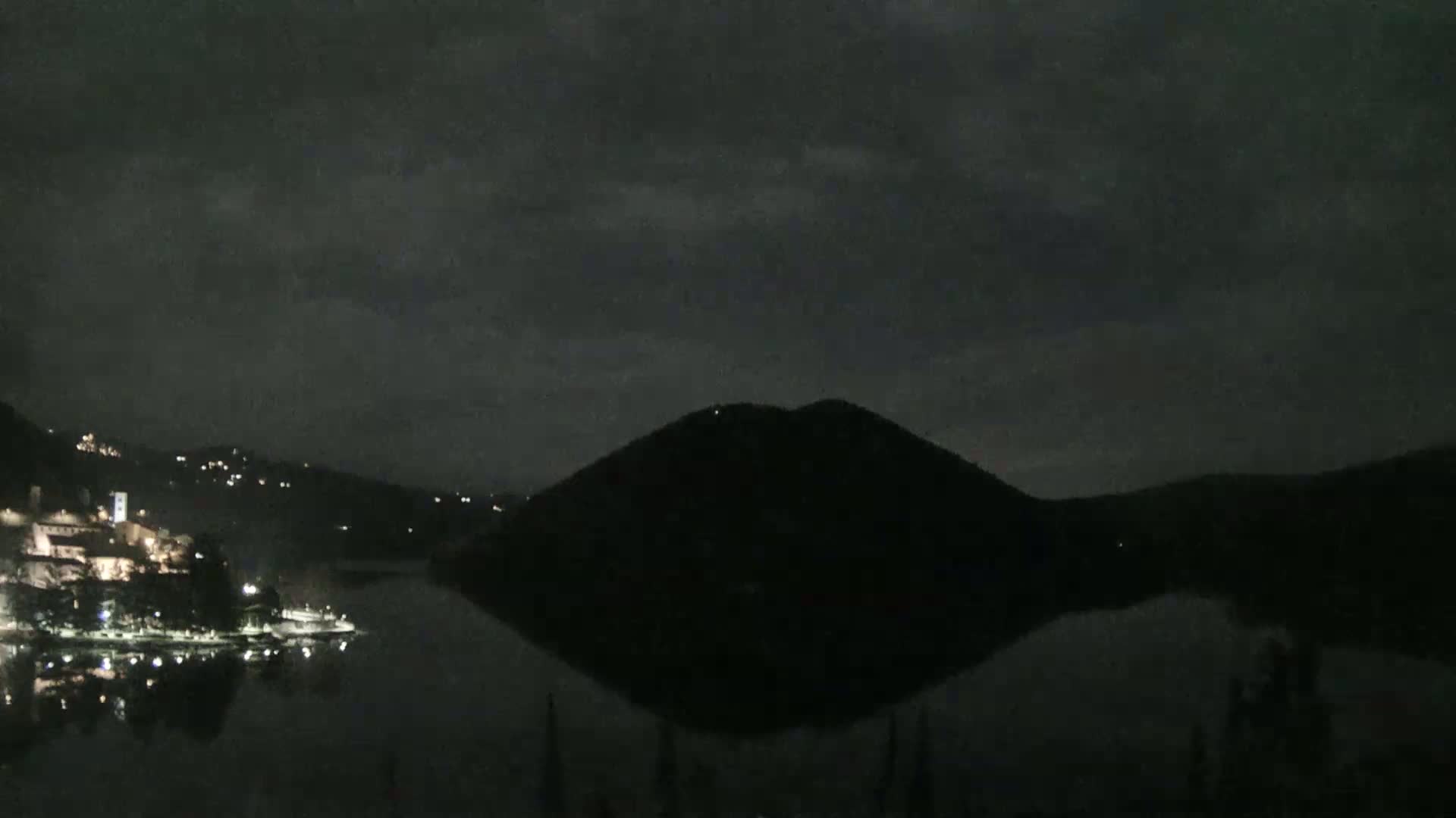 A dark, nighttime scene shows a body of water reflecting a hill and a small, lit town across the water under a cloudy sky.