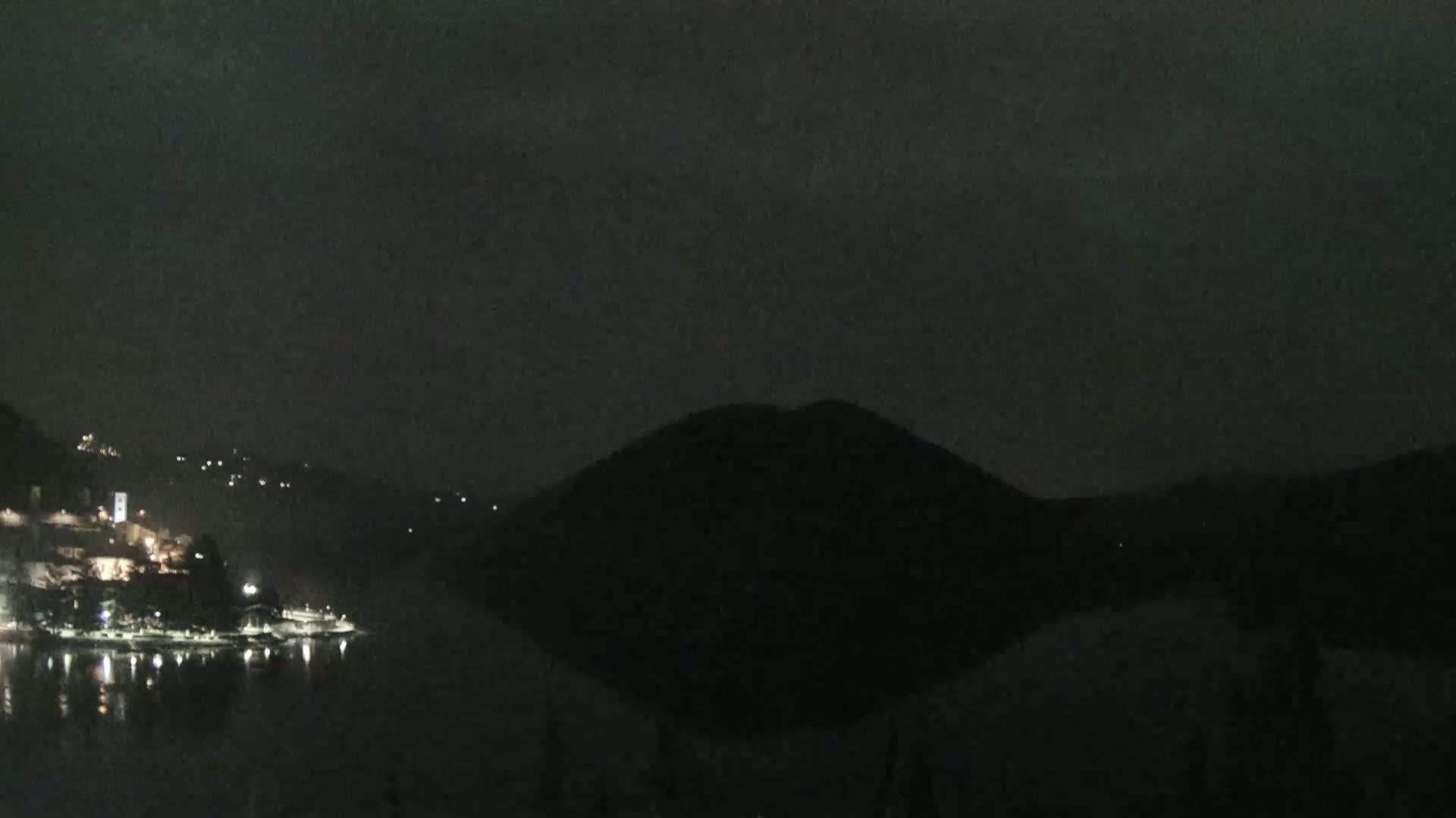 A dark, nighttime scene shows a body of water reflecting the lights of a town nestled on a hillside, with a large, silhouetted hill in the middle ground under an overcast sky.