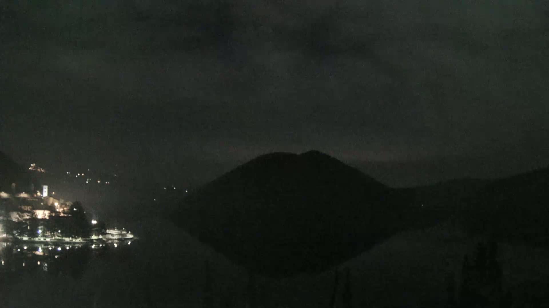 A dark, nighttime scene shows a body of water reflecting the lights of a small town nestled at the base of a silhouetted hill under a cloudy sky.